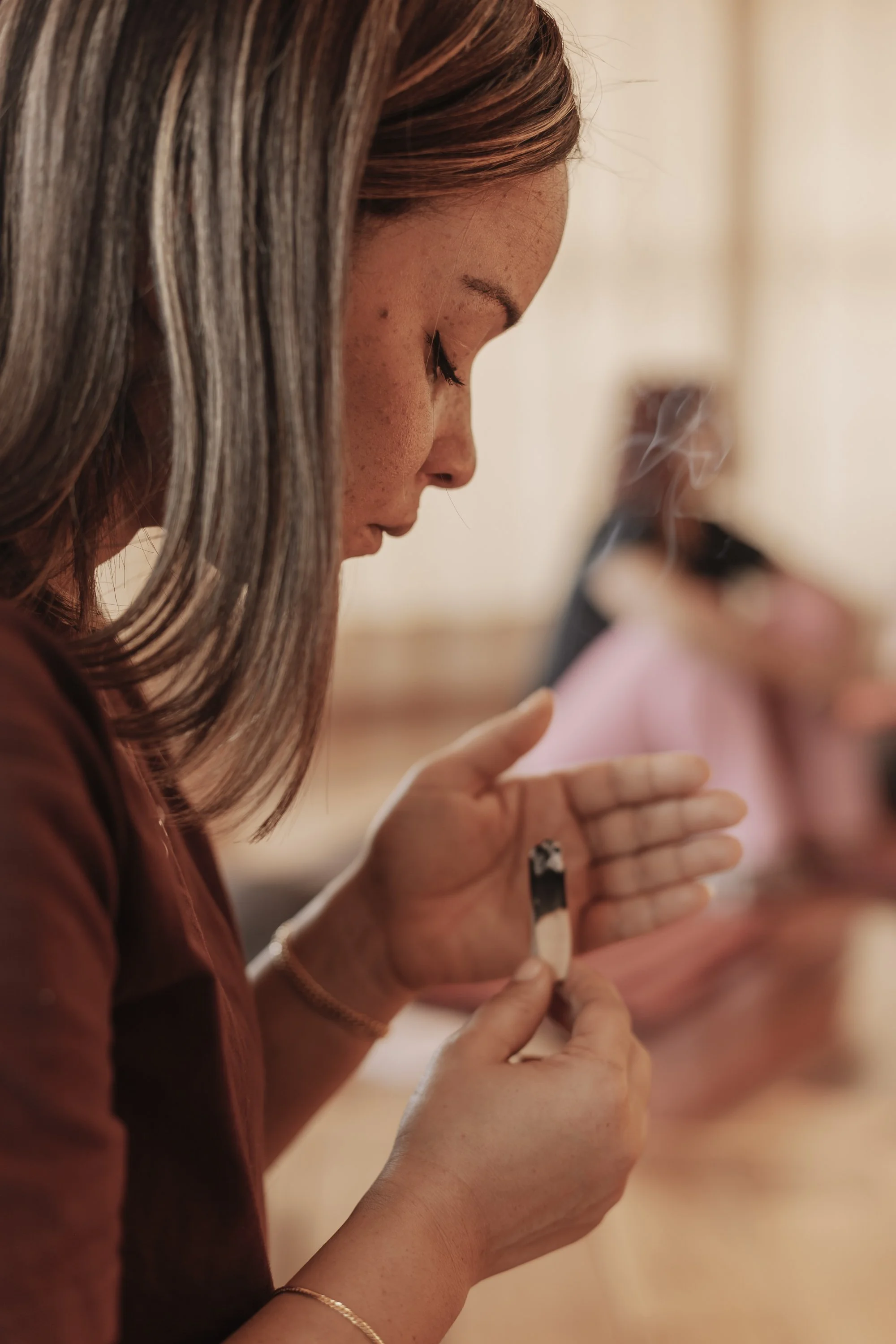 A woman with long, brown hair looking down as she holds a vape pen close to her lips, with smoke visible in the air.