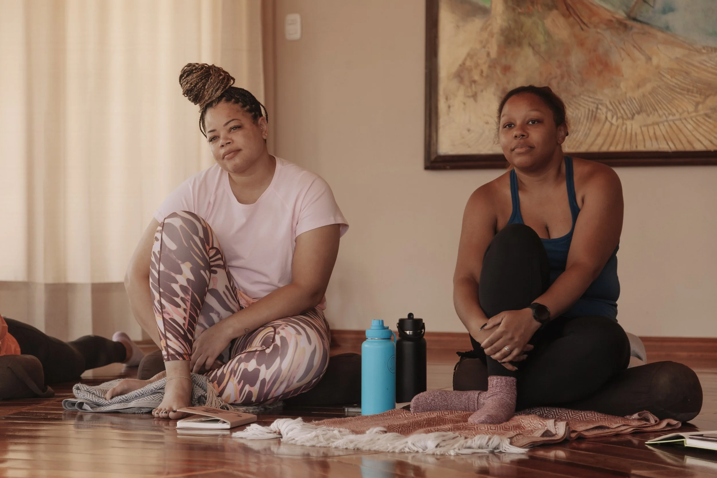 Two women sitting on the floor during a meditation class in a room with wooden flooring, water bottles in front, and a painting on the wall behind them.