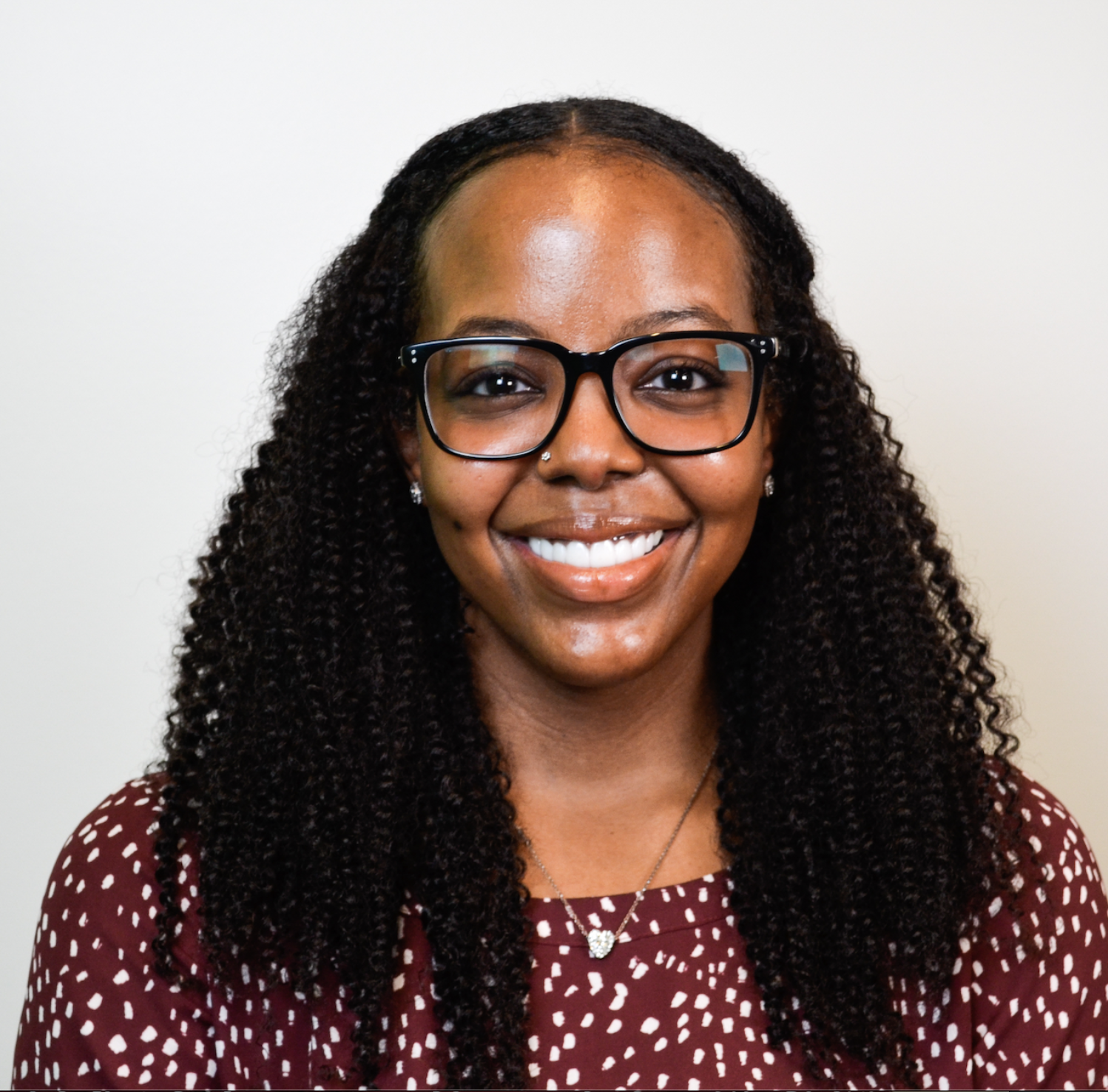 Smiling Black woman wearing black glasses, a maroon polka dot top, and a silver necklace with a heart pendant, against a plain white background.