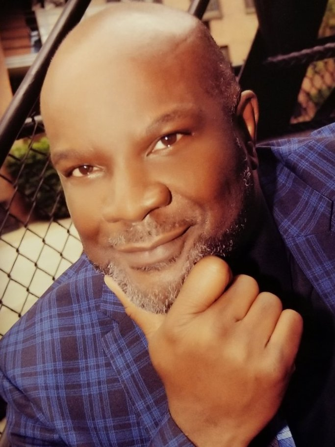 A man with a shaved head and gray beard smiling and resting his chin on his hand, dressed in a blue plaid jacket.