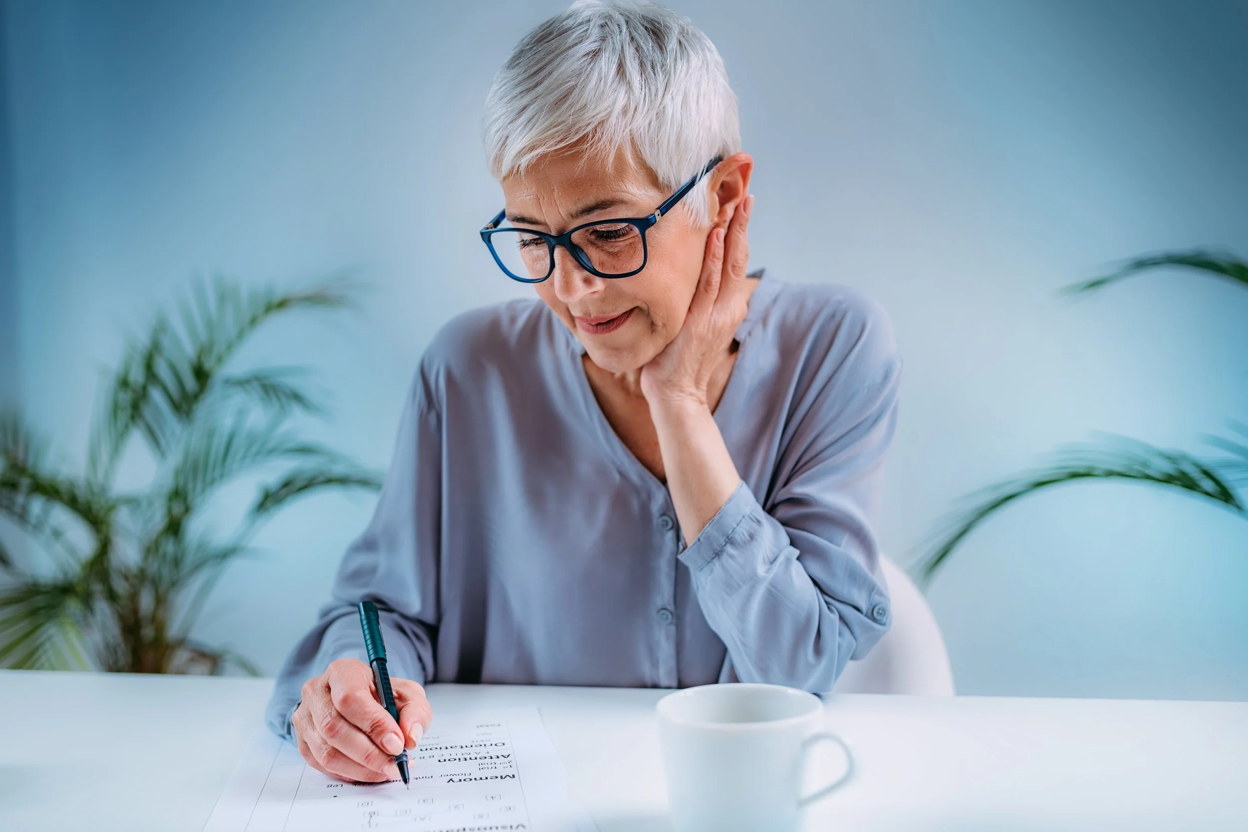 An elderly woman with short gray hair and glasses, sitting at a white table, working on a document with a black pen, with a white mug and plants in the background.