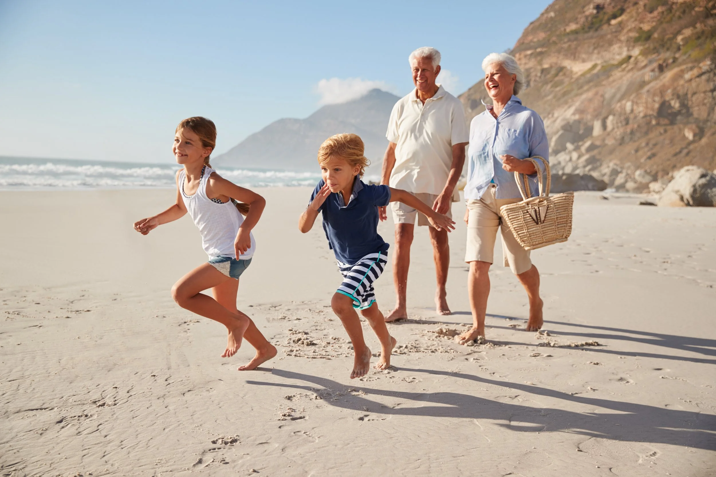 A multigenerational family of five enjoying a day at the beach, running along the sand with cliffs and the ocean in the background.