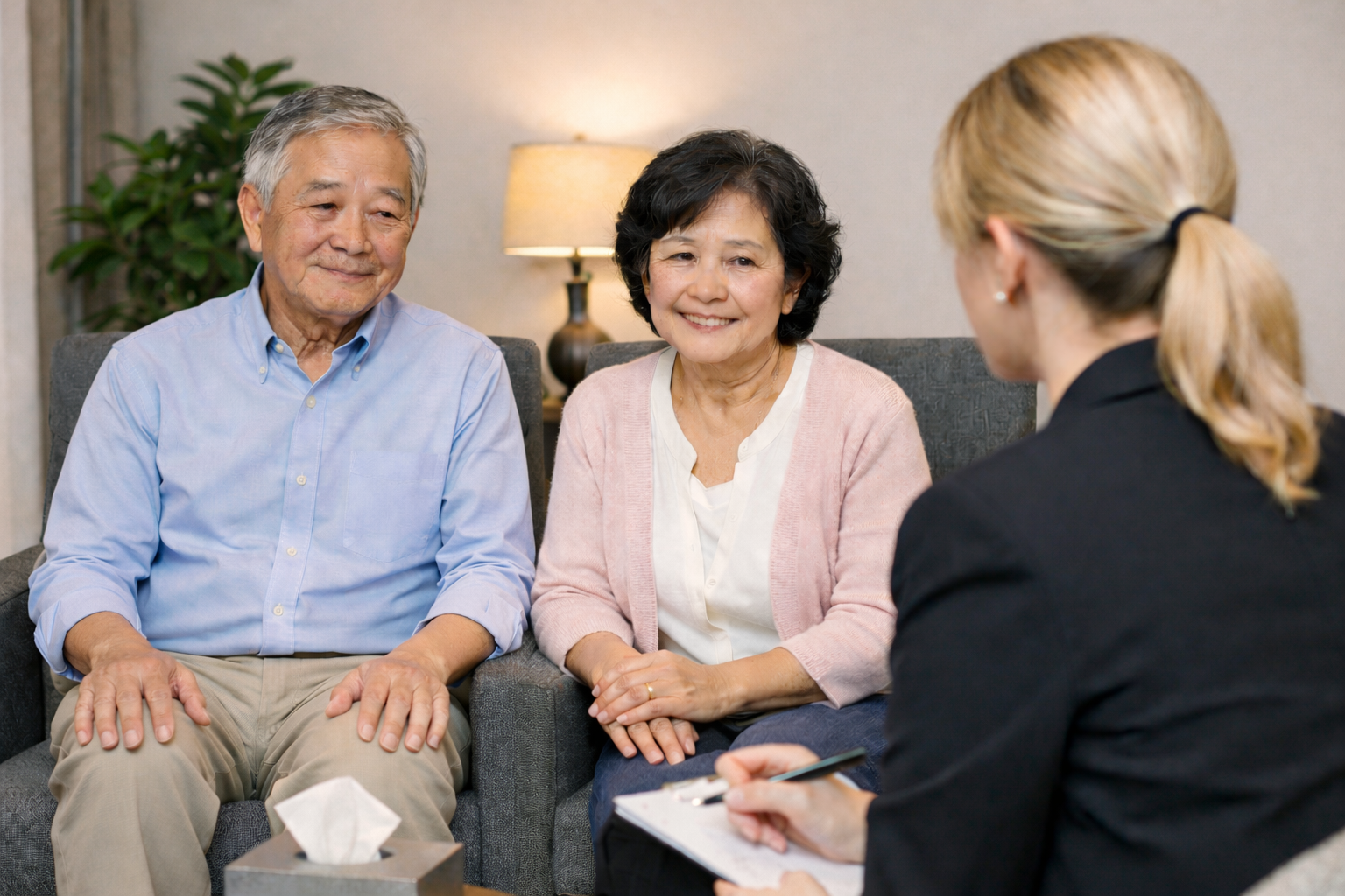 An elderly couple sitting on a sofa during a counseling session with a woman taking notes, in a cozy room with a lamp and a plant.