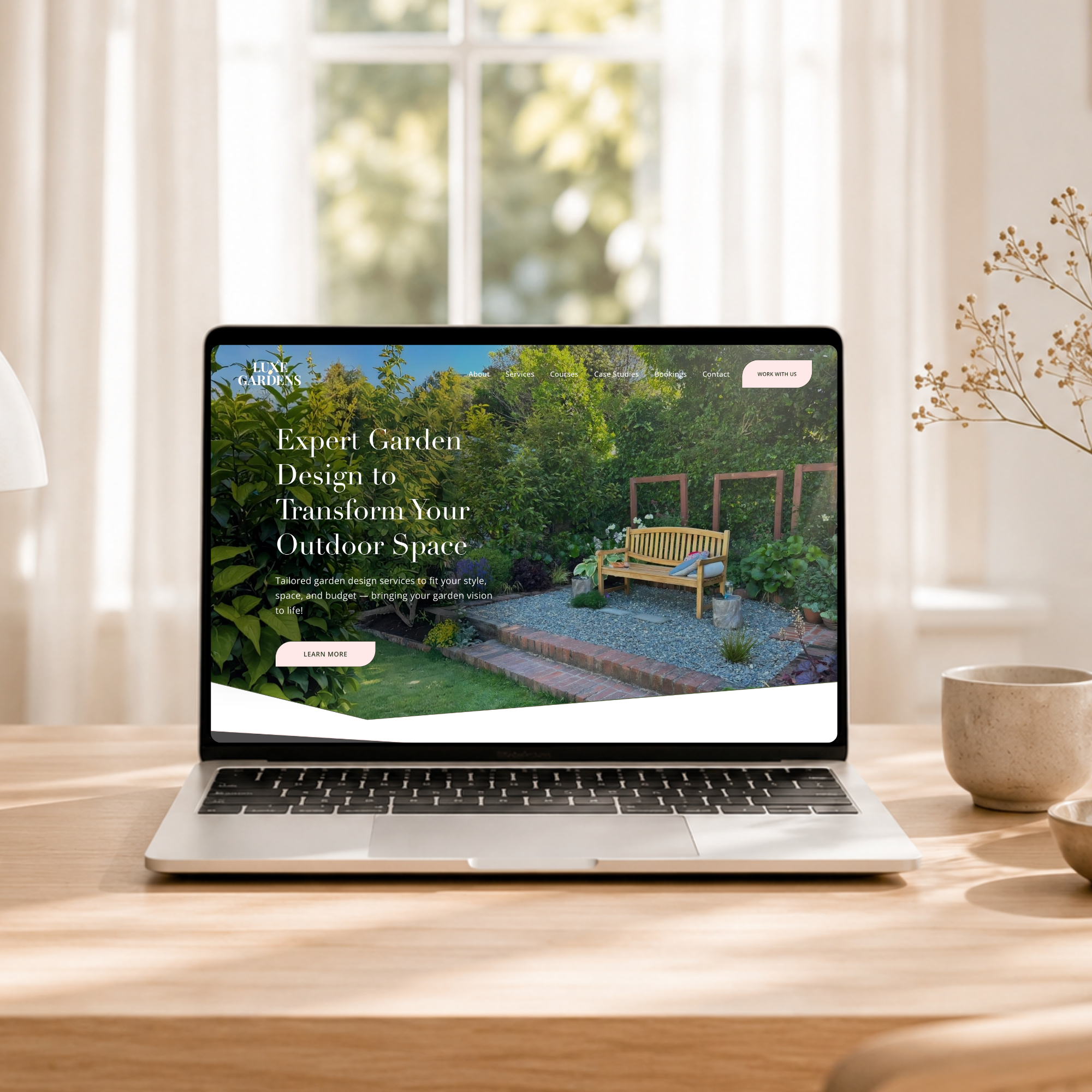 Open laptop on a wooden table in a light airy room displaying the hero section of the homepage of a garden design website with outdoor garden scene and seating area.