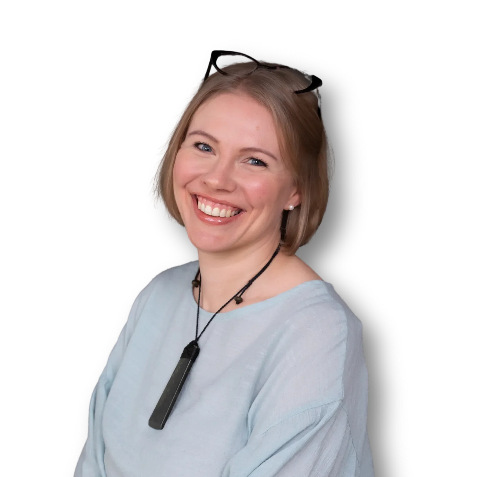 A smiling woman with short blonde hair, wearing glasses on her head, pearl earrings, a light gray shirt, and a pounamu necklace with a rectangular greenstone pendant, against a black background.
