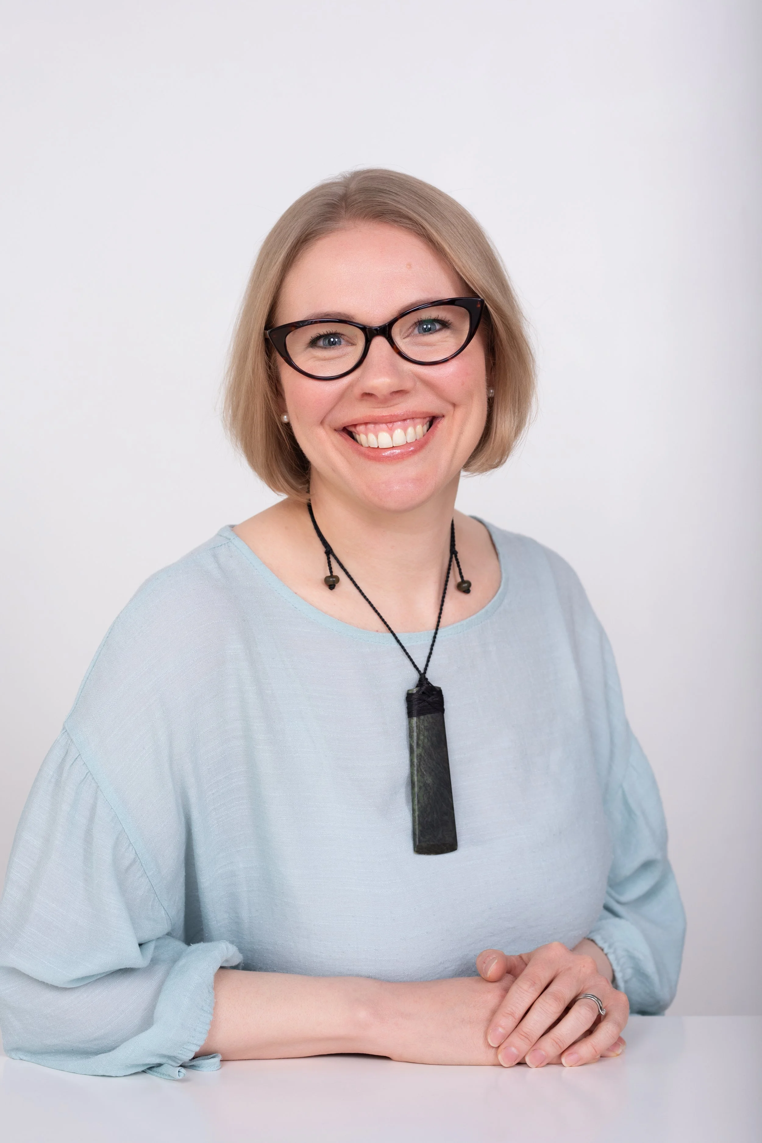 A woman with short blonde hair, wearing black glasses and a light blue top, smiling at the camera with hands resting on a white table in front of her.