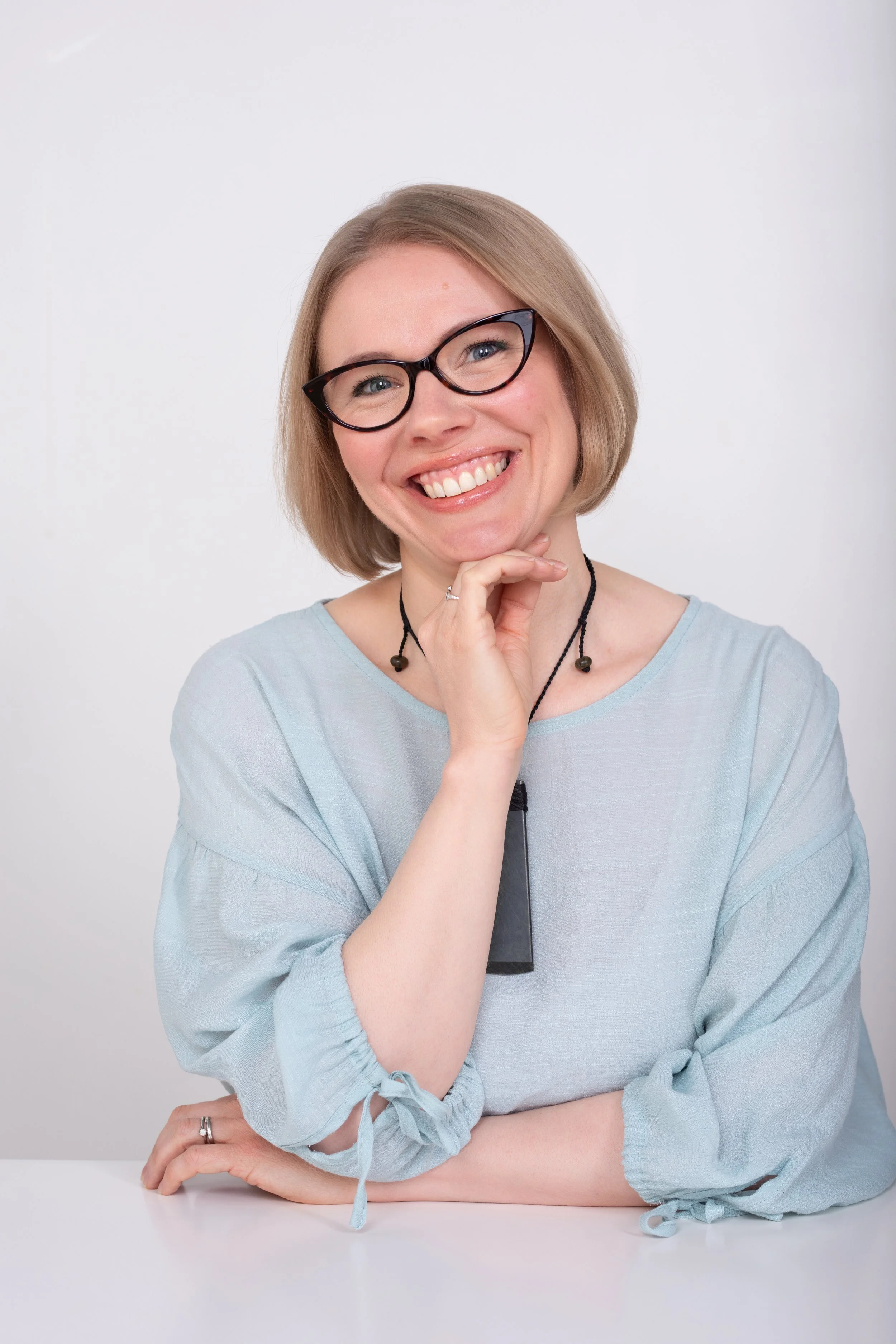 A woman with light brown hair, black glasses, and a bright smile. She's wearing a light blue top and a greenstone pendant, sitting at a white table against a plain white background.