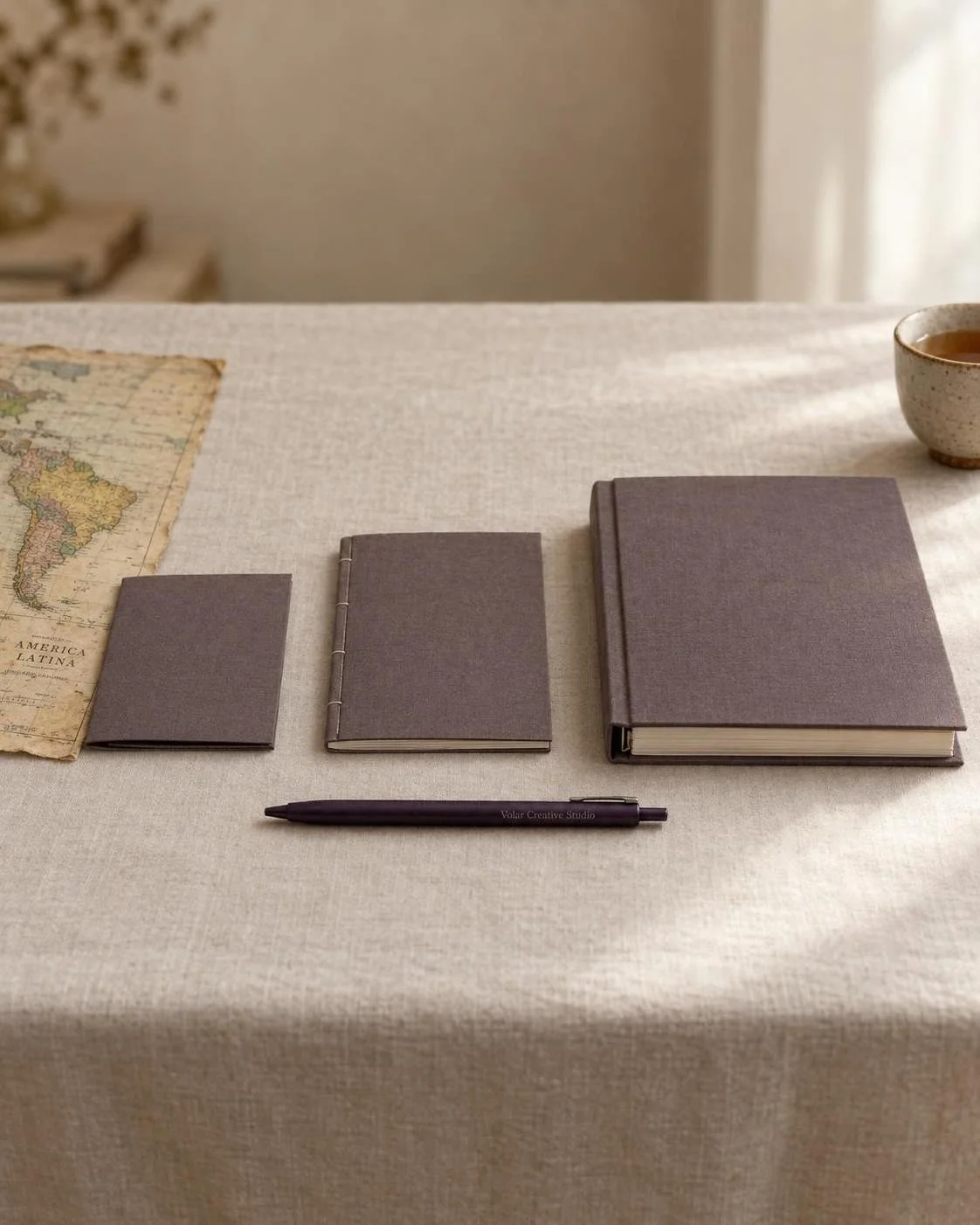 Three brown hardcover notebooks, a pen, a vintage Latin America map, and a cup of tea on a linen-covered table