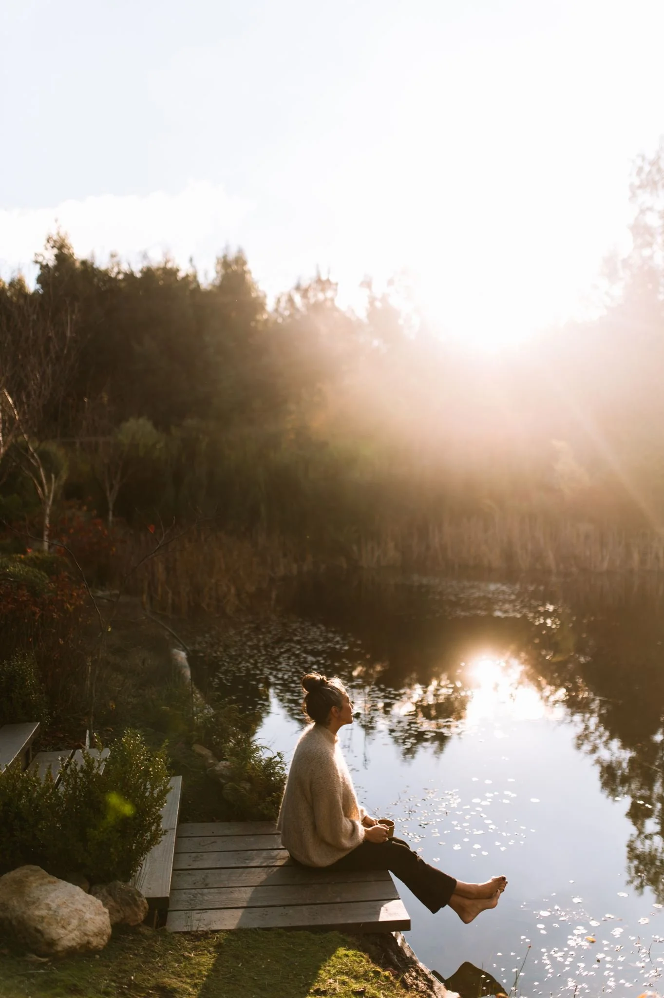 A woman sitting on a wooden dock by a lake during sunset, holding a cup, with her feet dipped in the water, surrounded by trees and nature.