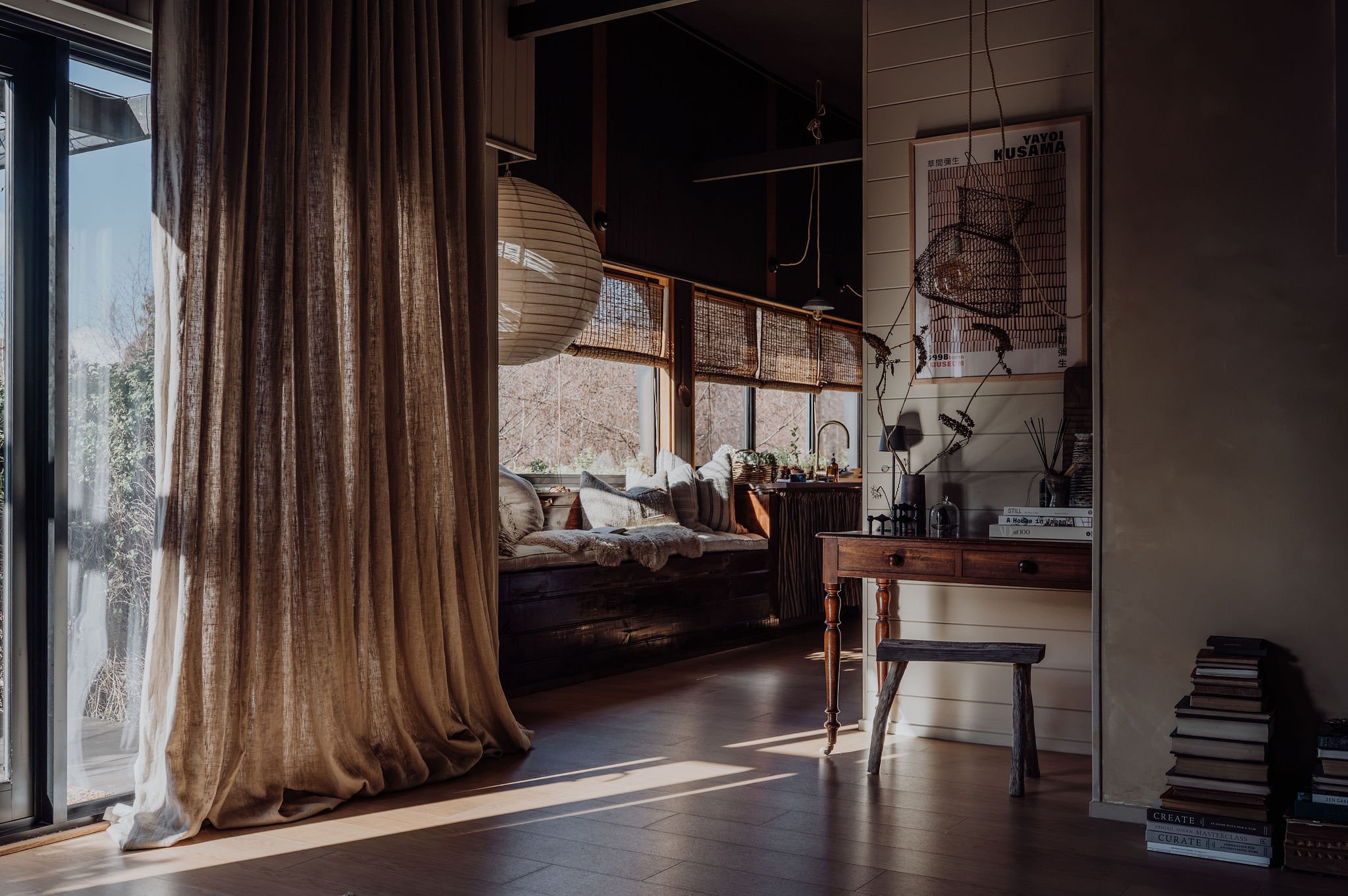 Interior of a cozy living room with large windows, beige curtains, a built-in window seat with cushions, a round paper lantern hanging from the ceiling, a small wooden table, potted plants, and a framed poster on the wall.