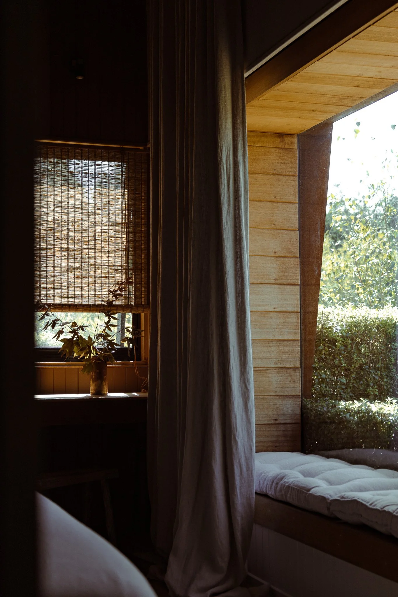 A cozy corner of a wooden room with natural light, featuring a window with bamboo blinds, a plant in a vase on the windowsill, and a cushioned seating area near large windows showing greenery outside.