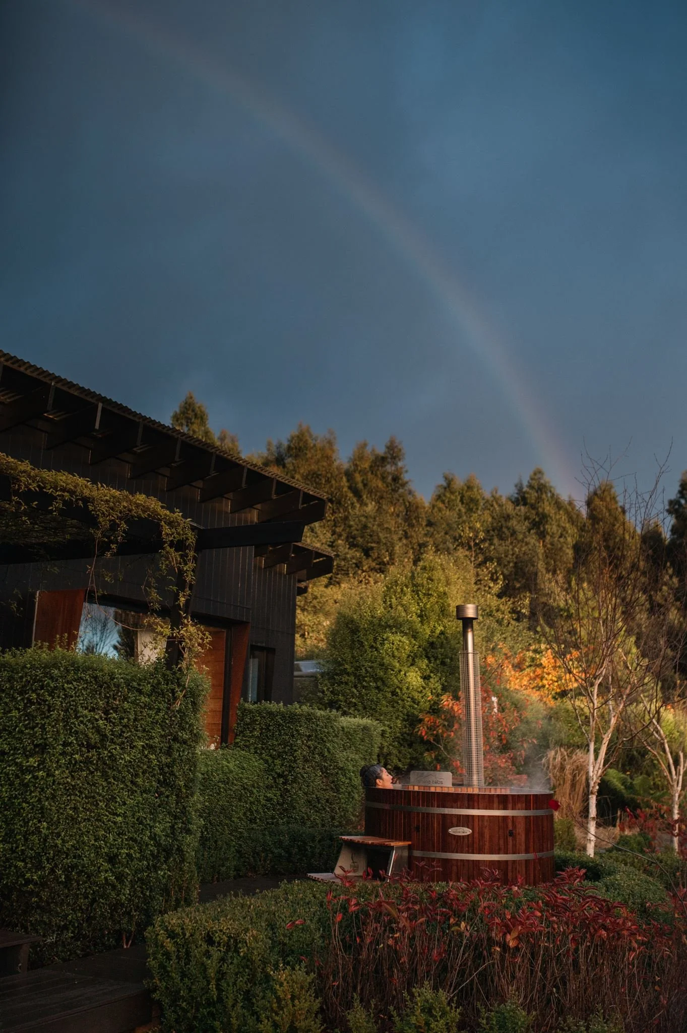 A person relaxing in a hot tub outdoors surrounded by lush greenery and trees during sunset, with a modern house visible to the left and a rainbow in the sky.