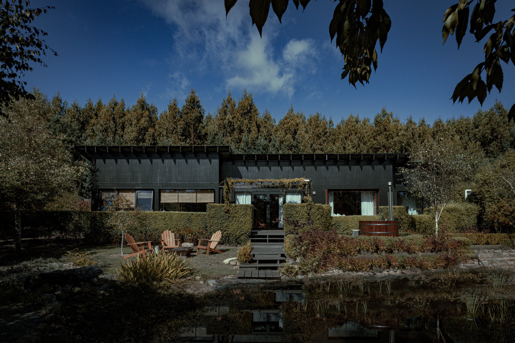 Modern black house with large windows, surrounded by trees and bushes, with a grassy yard and Adirondack chairs by a pond, under a blue sky with some clouds.