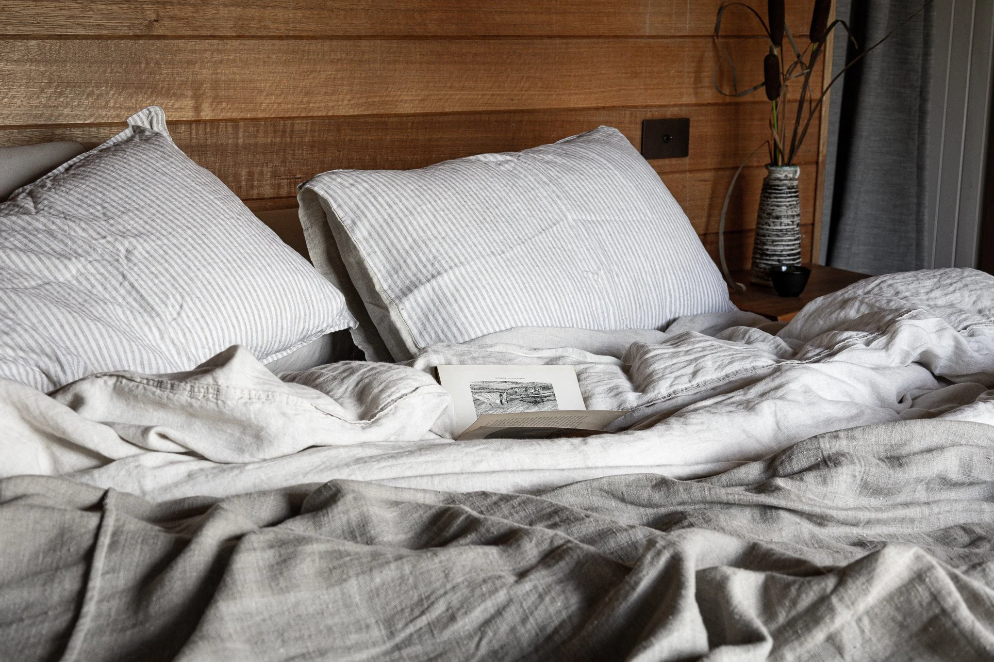 A neatly made bed with white and beige striped pillows, a book and a photograph on top, and a wooden headboard. There is a bedside table with a decorative vase and some plants in the background.