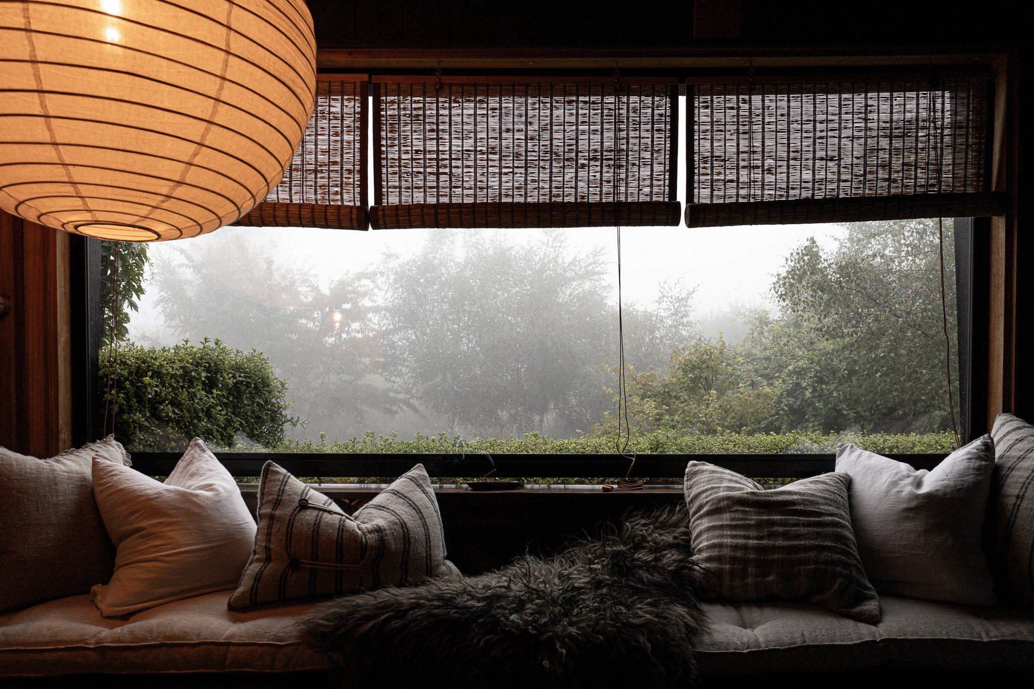 Indoor seating area with a curtain of pillows in front of a large window showing foggy trees outside, and a large round paper lantern hanging from the ceiling.