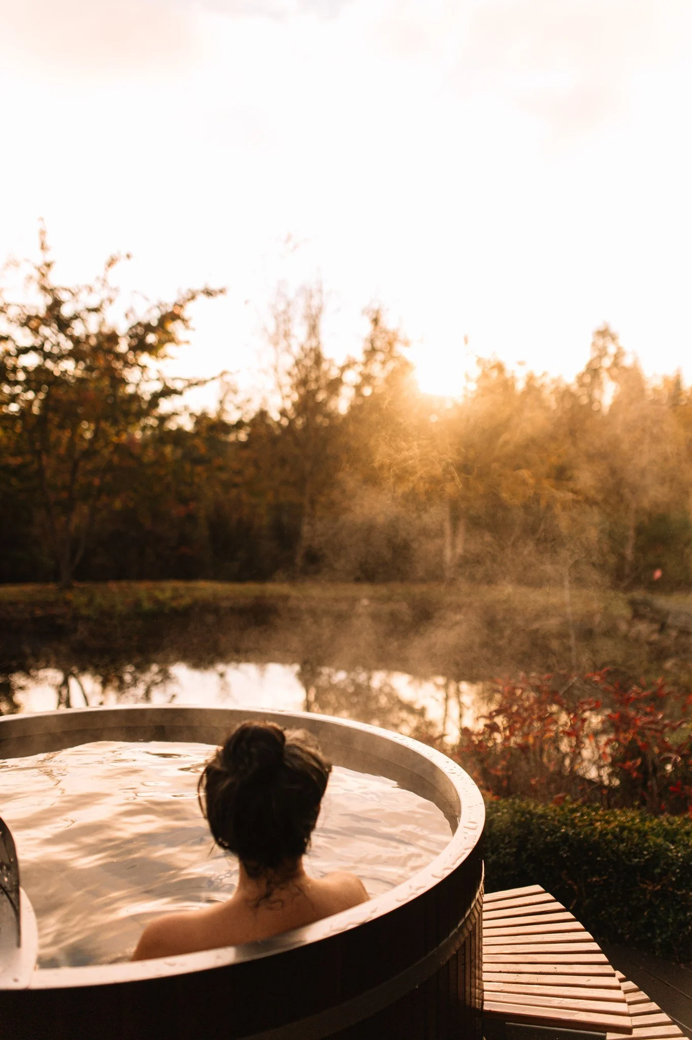 Person relaxing in an outdoor hot tub by a lake during sunset, with trees and a calm water reflection in the background.