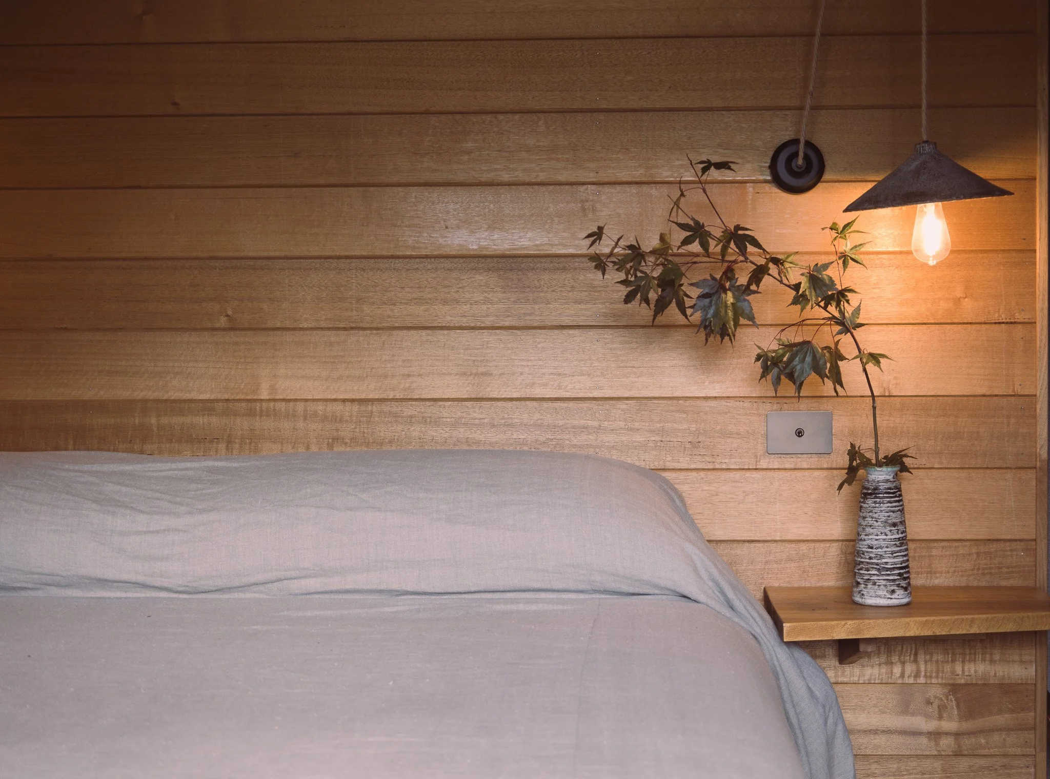 A bedroom with a bed covered in a light-colored sheet, against a wooden paneled wall. There is a wooden nightstand with a vase holding a leafy branch and a hanging light with an exposed bulb.