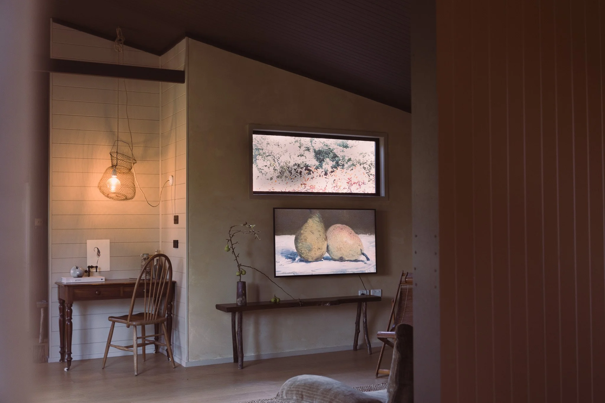 Living room with a small wooden desk, wooden chair, a hanging light fixture, a long wooden bench with a potted branch, a window, and a wall-mounted TV displaying pears, with vertical striped wallpaper.