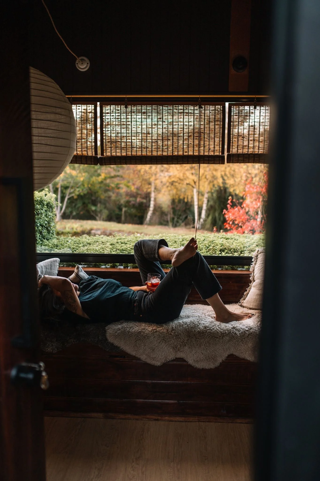 A person reclining on a cushioned window seat, holding a glass of drink, with a scenic view of colorful autumn trees outside the window. The scene is viewed from outside a doorway.