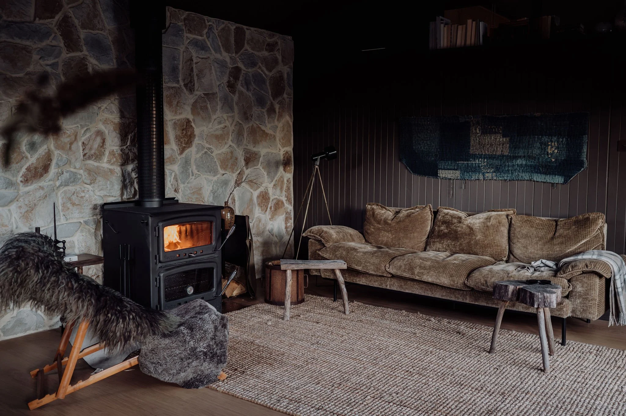 Living room with a stone fireplace, a wood stove, and a brown sofa with multiple pillows. A map or textile hangs on a dark wood paneled wall, with books on a shelf above. A tripod floor lamp and small wooden table are also present.