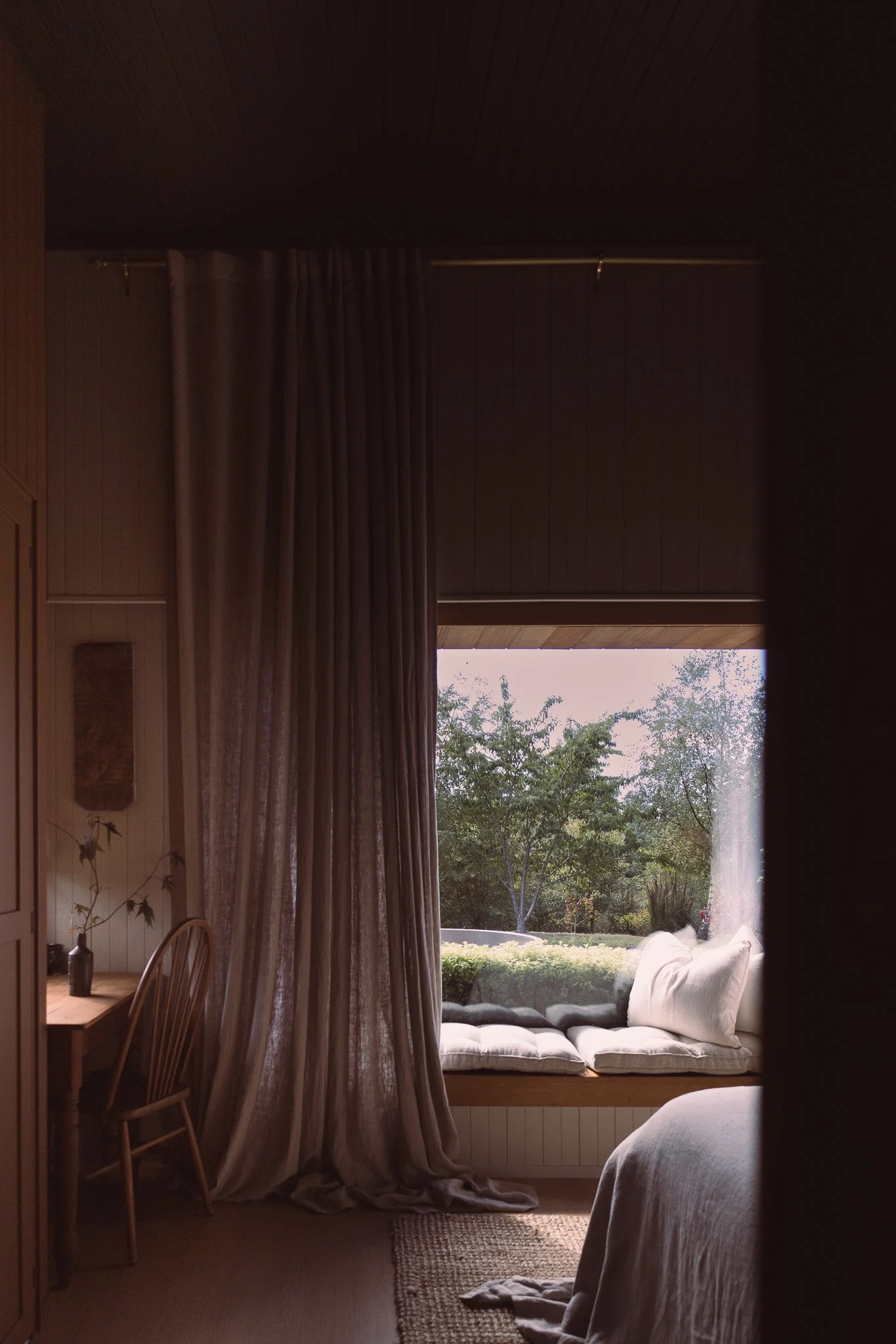 Interior view of a cozy room with a window seat, beige curtains, a wooden desk and chair, and a bed with white bedding. Outside the window, trees are visible.