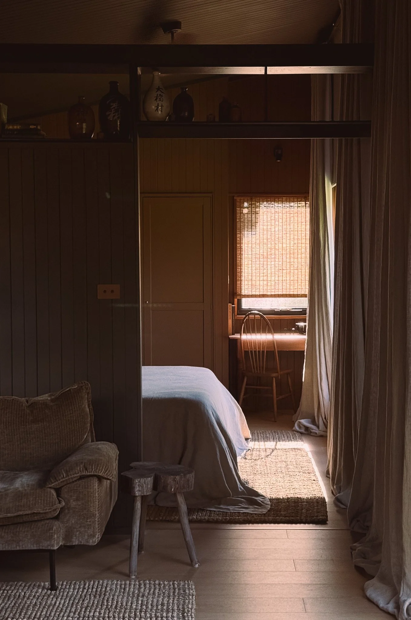 Interior of a cozy room with a bed near a window, a wooden desk with a chair, and a sofa, decorated in warm earthy tones with natural light streaming in.