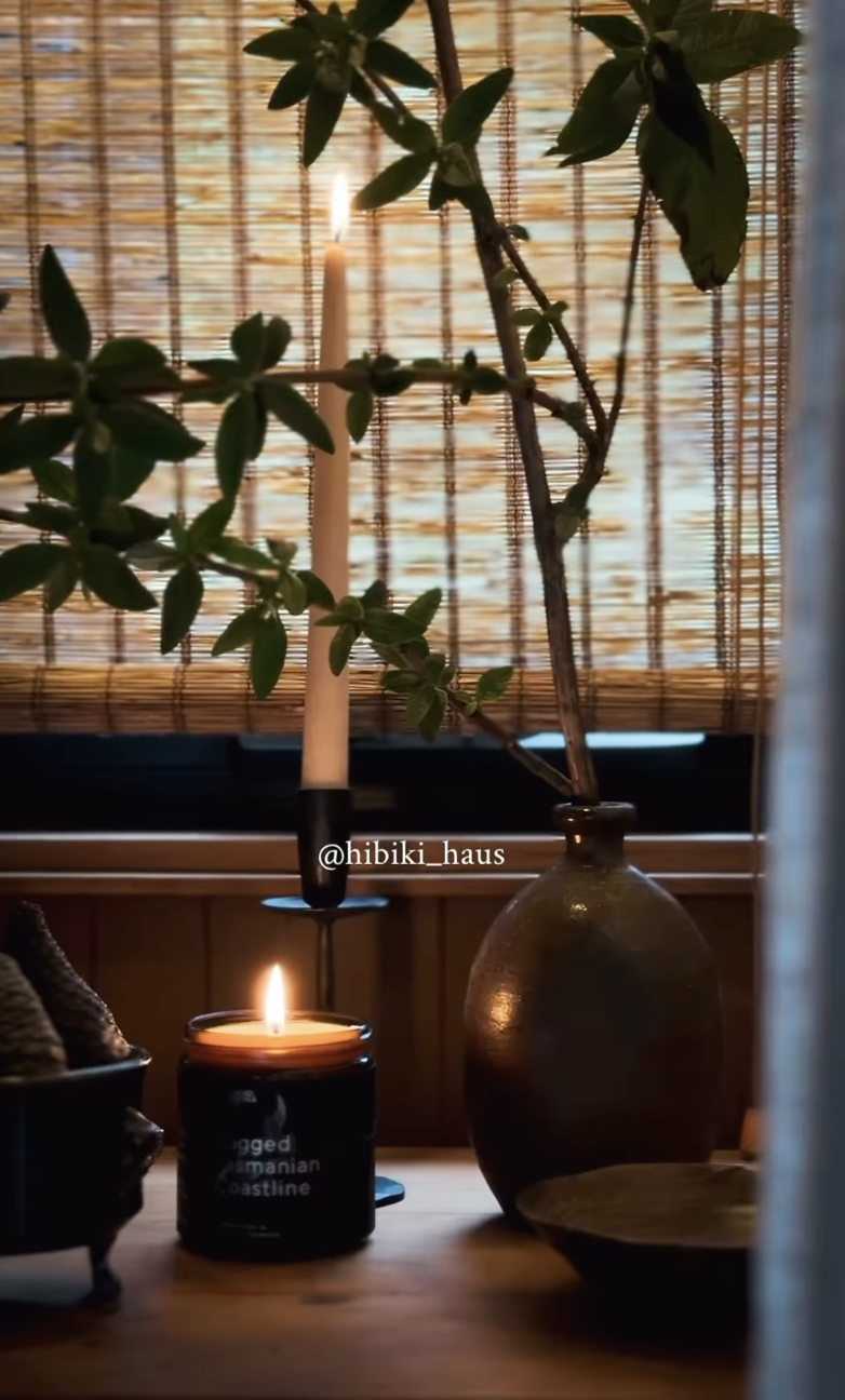 A lit candle in a black container, a large glass vase with green leafy branches, a bamboo curtain in the background, and a small dish on a wooden surface.