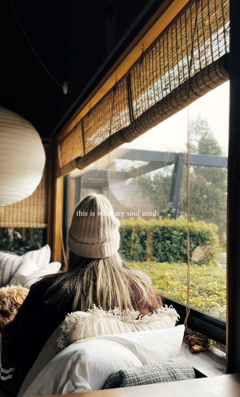 A woman with long hair wearing a beige knit beanie sitting on a cozy couch, looking out a large window with greenery outside, with the text 'this is what my soul needs' overlaid.