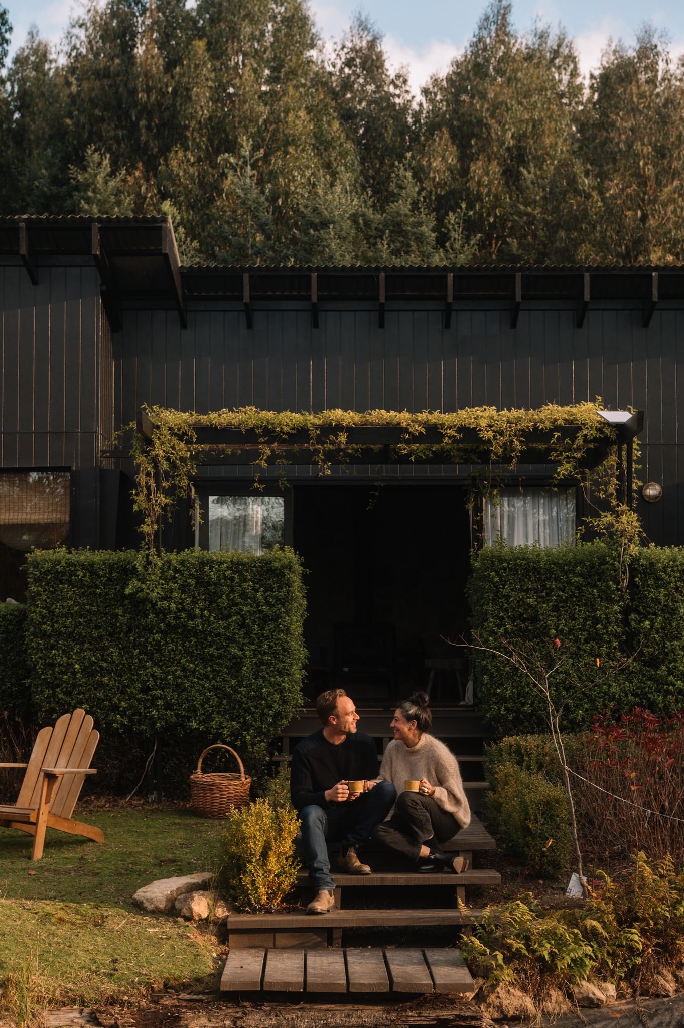 A couple enjoying coffee on a wooden step outside their house, surrounded by lush greenery and garden furniture.