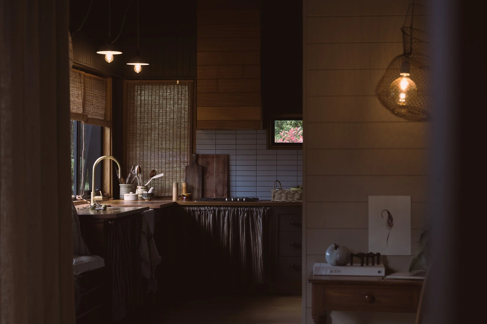 A cozy kitchen interior with warm lighting, wooden cabinets, a window with bamboo shades, a sink, and various kitchen utensils.