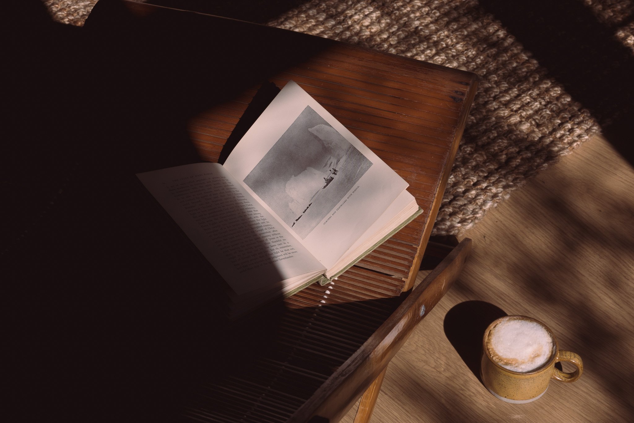 An open book with a black-and-white photograph on one page, resting on a wooden table, with a yellow cup of frothy coffee nearby, and a textured woven rug in the background.