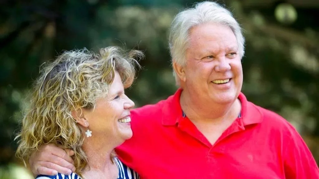 A smiling woman with curly blonde hair and a man with short gray hair, both smiling and enjoying each other's company outdoors.