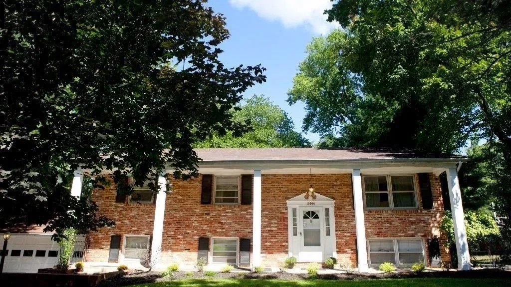 Front view of a brick house with white columns, a white front door, and greenery including trees and shrubs in a suburban neighborhood.