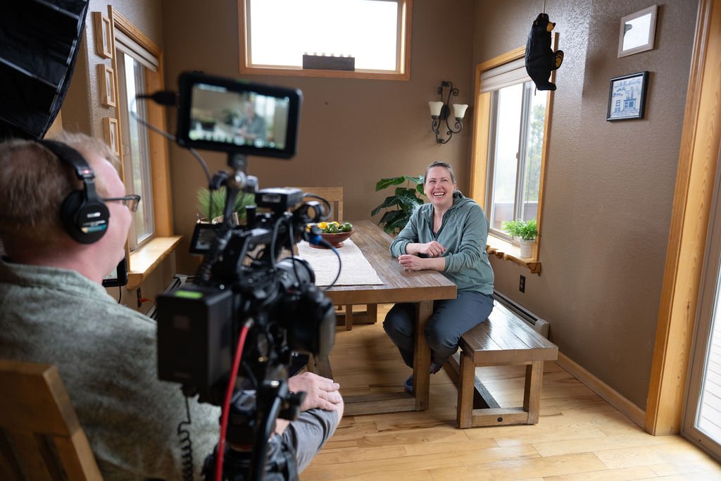 A woman sitting at a wooden dining table, smiling and talking, while being filmed by a person using a professional camera and wearing headphones in a cozy, well-lit kitchen with large windows and houseplants.