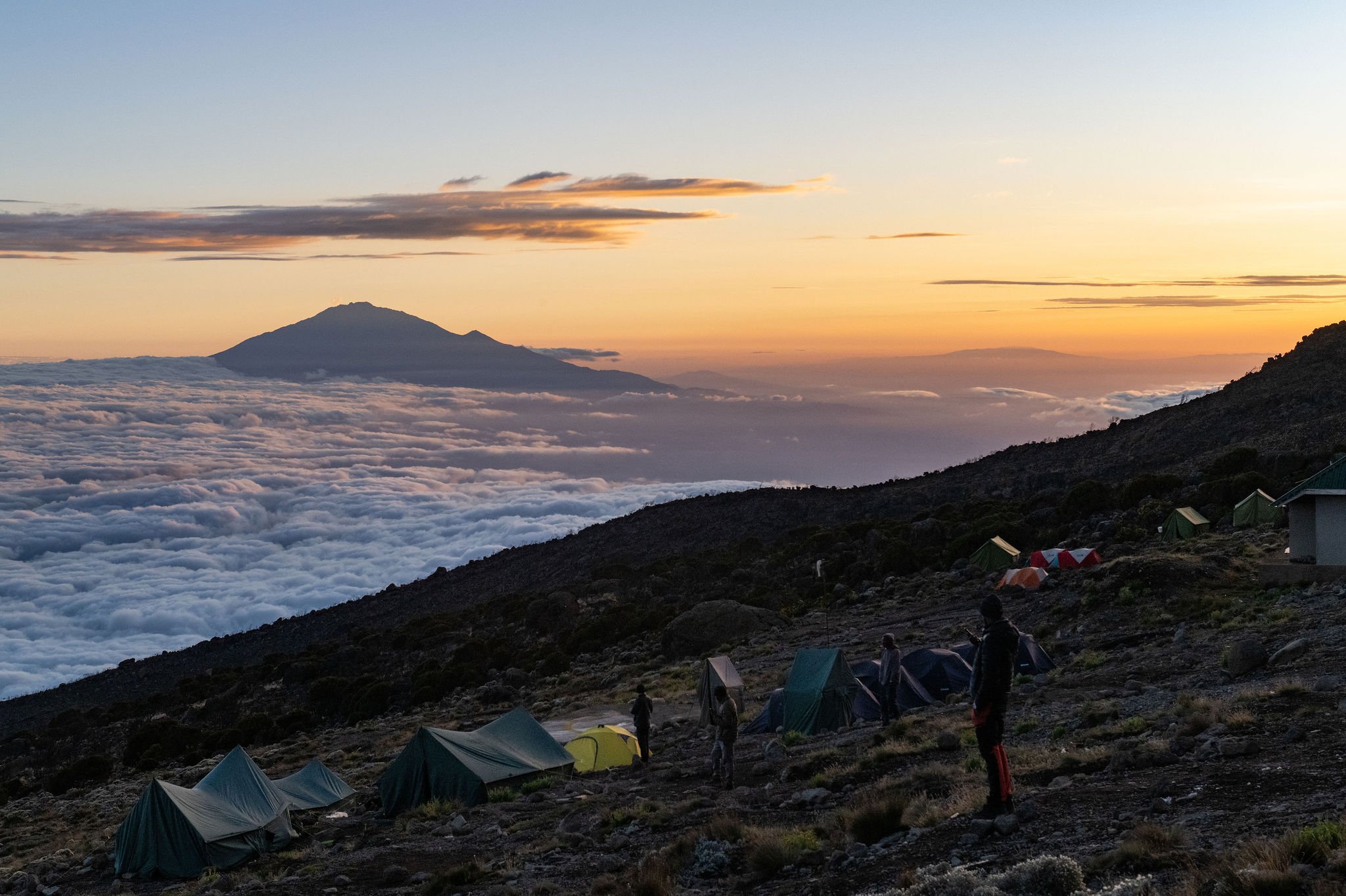 Campgrounds on a mountain slope at sunset with a mountain in the background and clouds below, and several people walking around the tents.