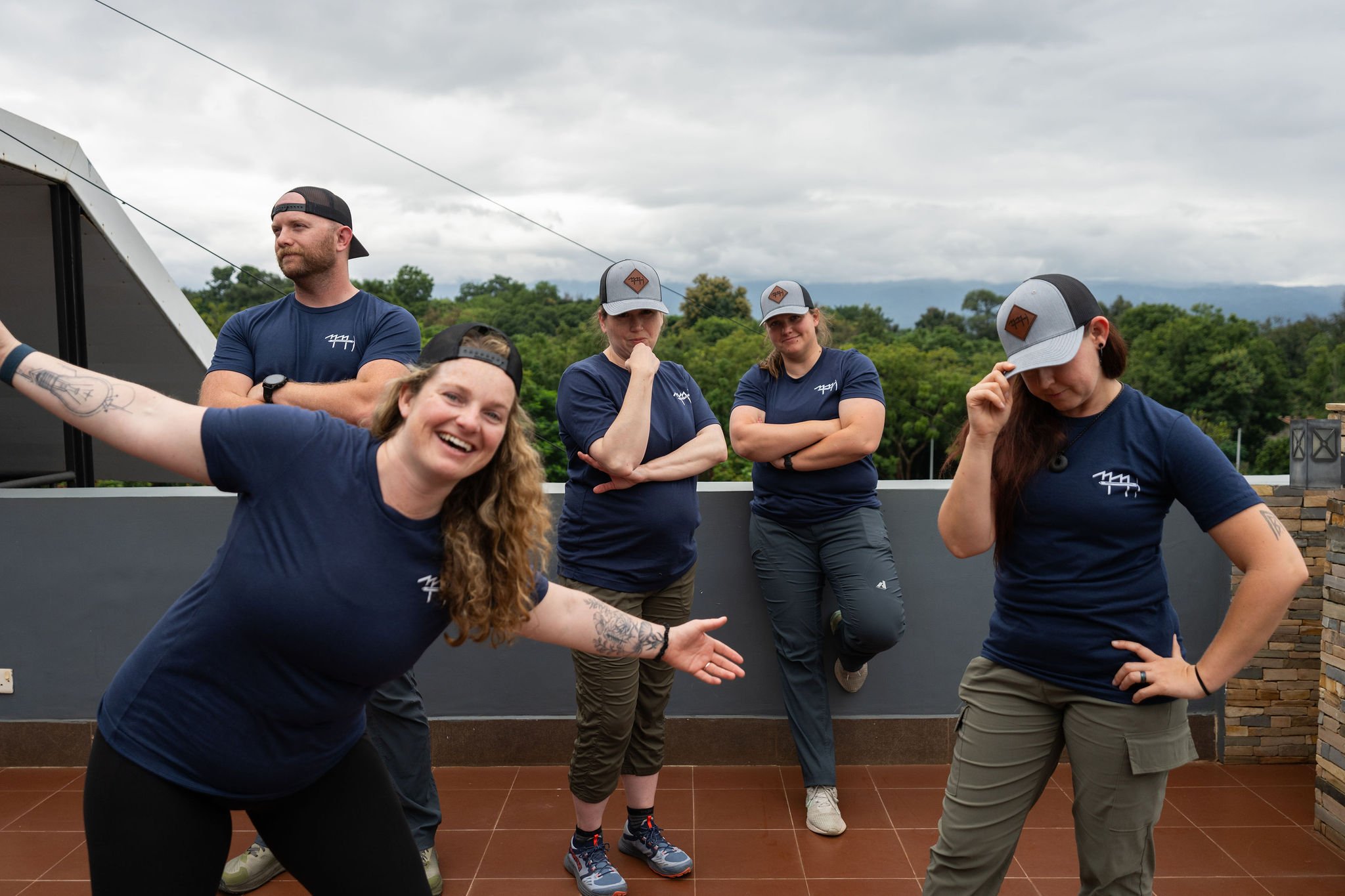 Five people on a rooftop patio, four women and one man, wearing matching navy blue shirts and caps, with a backdrop of green trees and cloudy sky.
