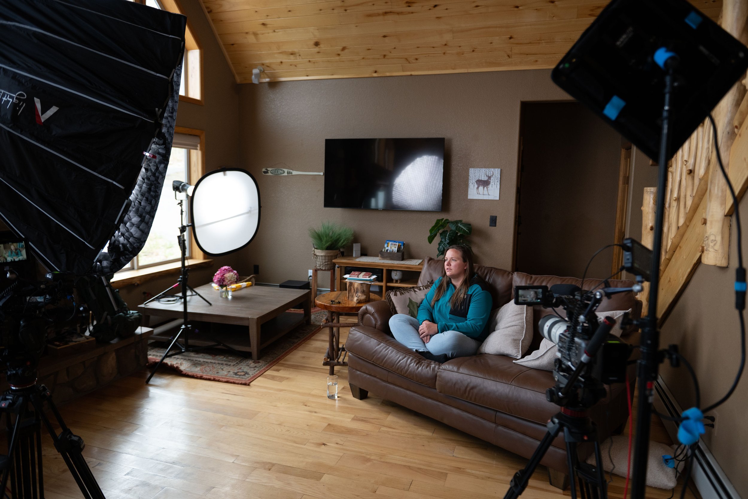 A woman sitting on a brown leather couch in a cozy living room with filming equipment around her, including lights and cameras, during a recording or interview session.