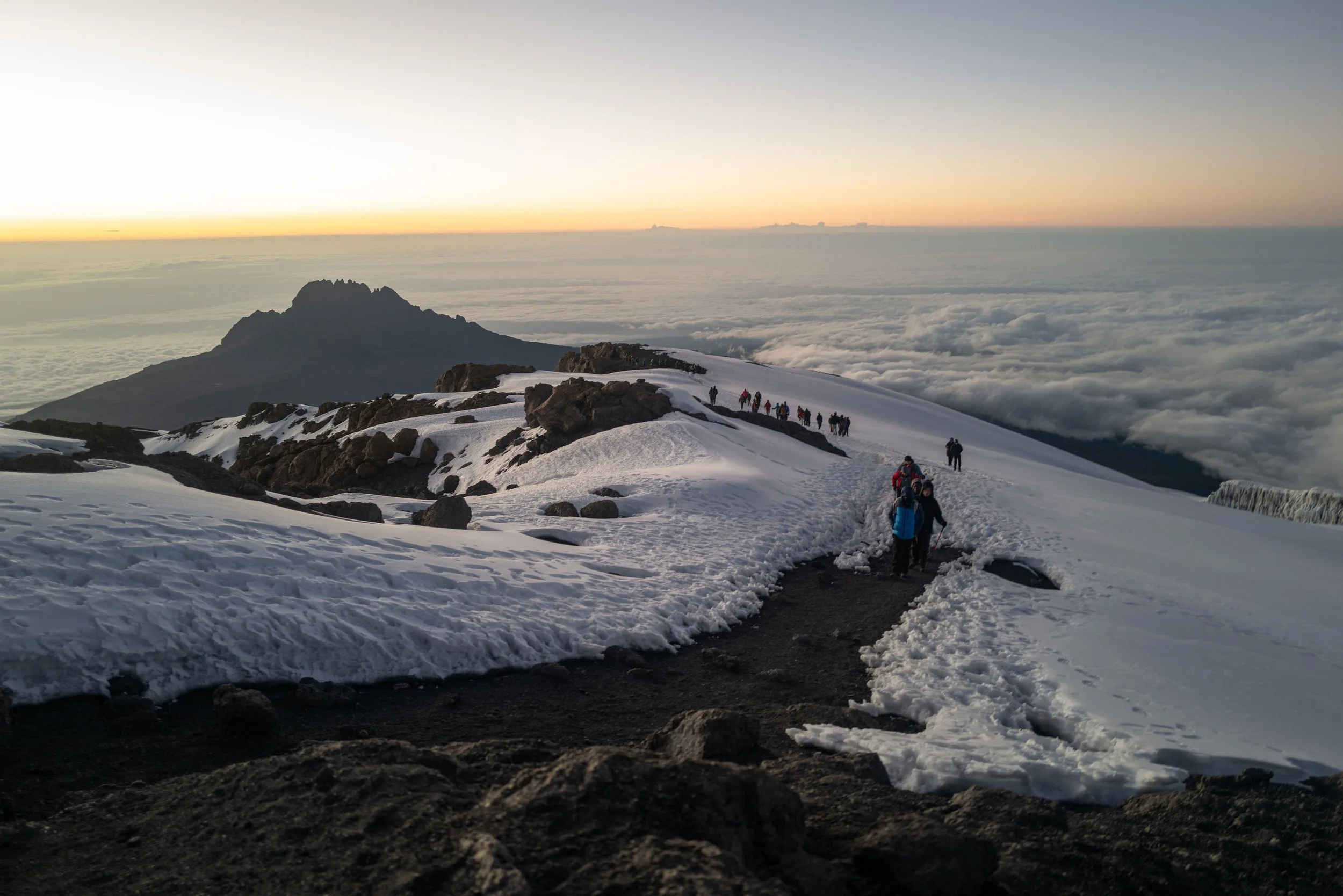 Group of hikers walking along a snowy mountain trail during sunset, with clouds below and mountains in the background.