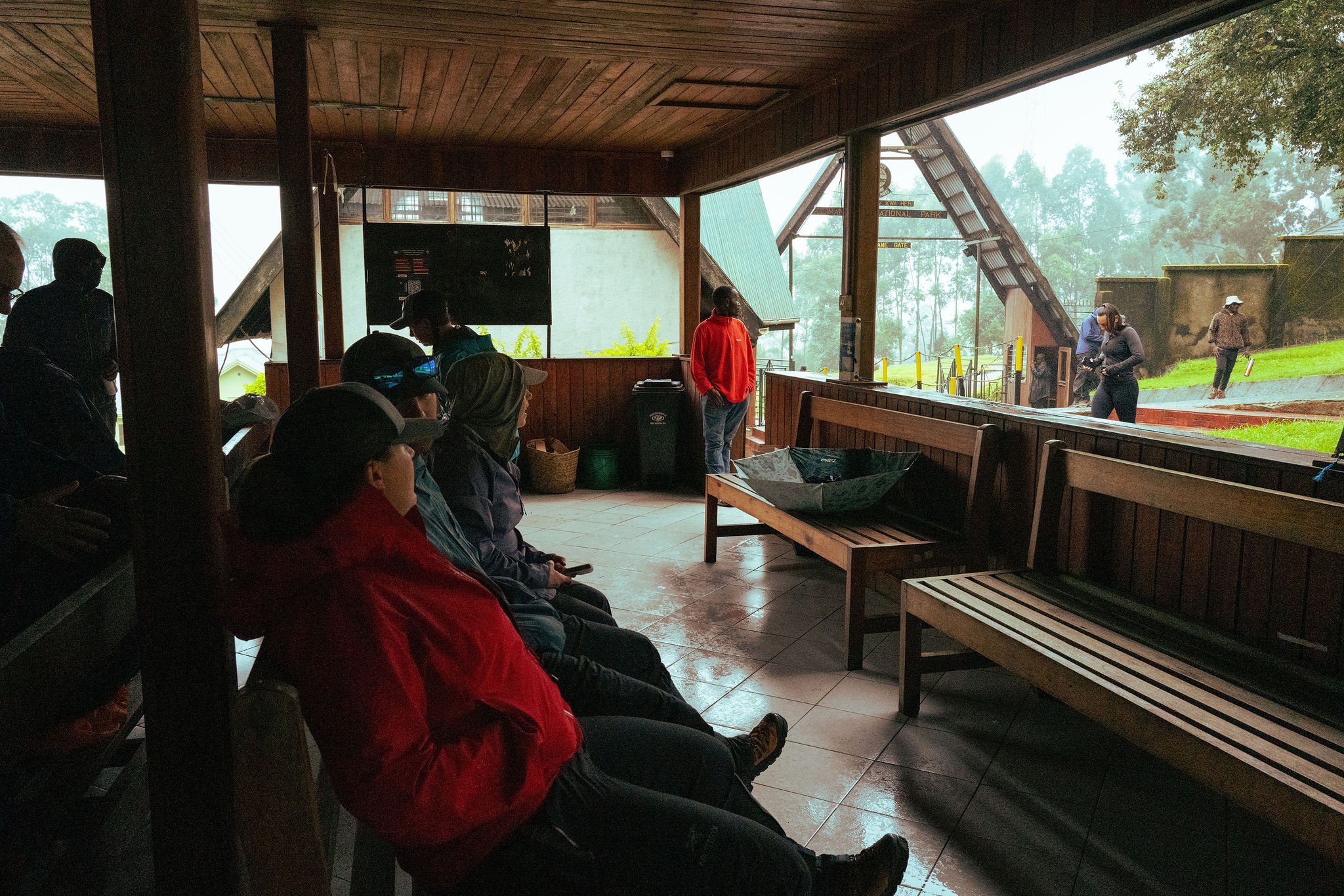 People seated inside a sheltered viewing area at a park, looking toward a hillside with trees and visitors near a large, open gate.