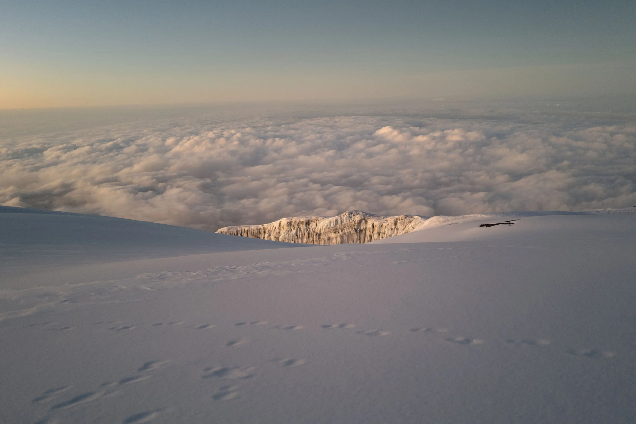 Snow-covered mountain slope with animal tracks, clouds below, and a distant snow-capped ridge at sunrise.