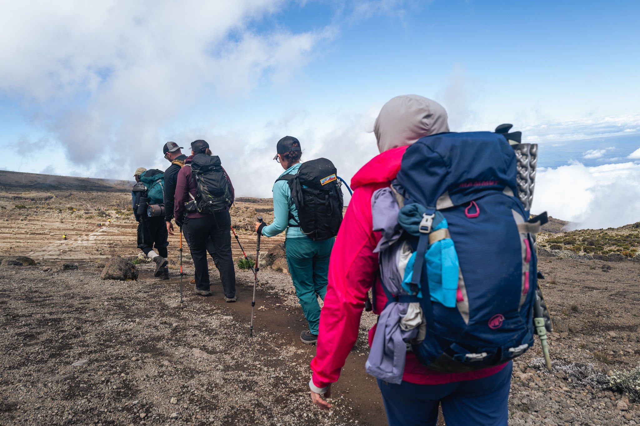 Group of hikers walking in a line on a trail on a mountainous landscape with clouds in the sky.