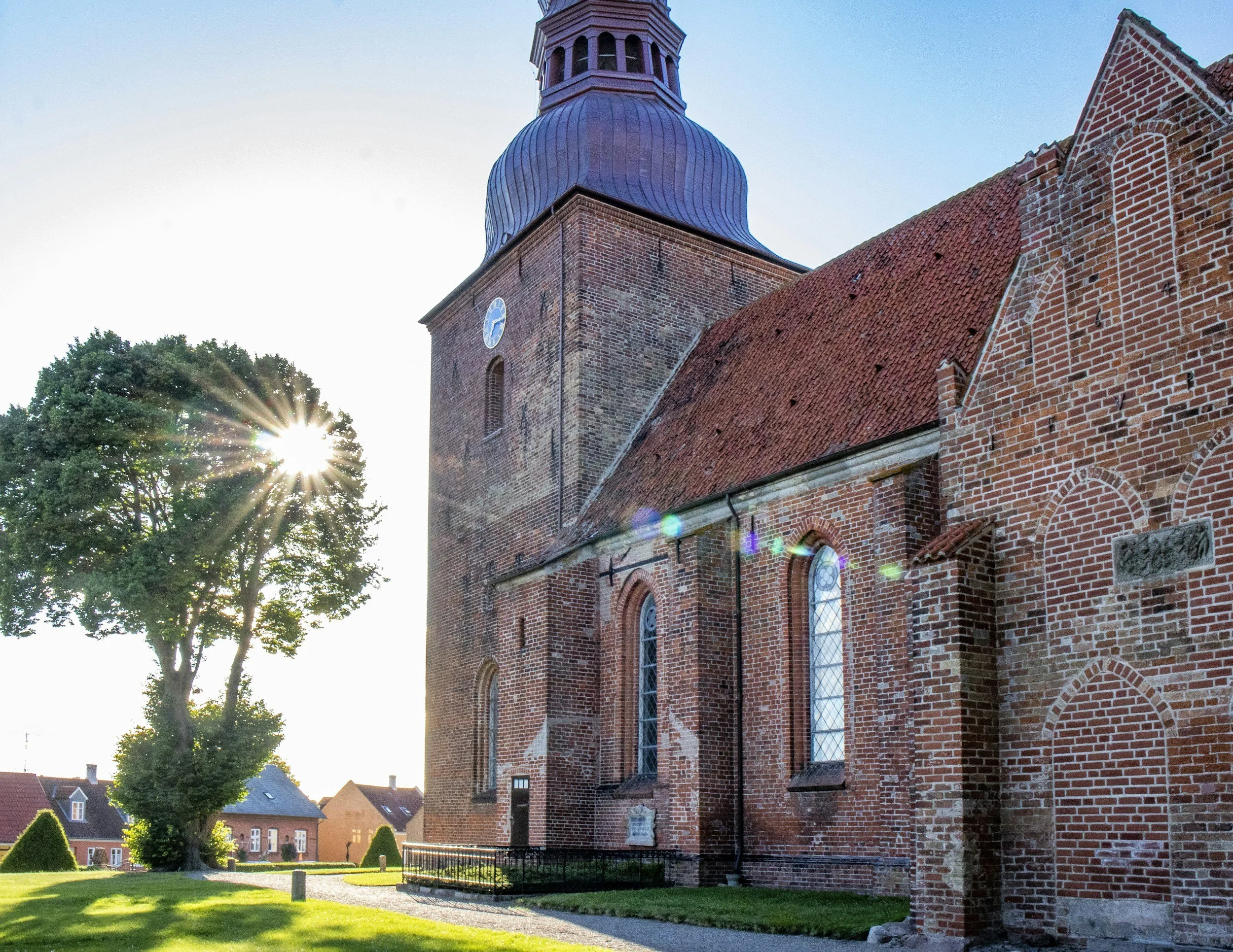 A brick church with a blue bell tower and arched windows, sunlight shining through a large tree nearby, with small houses in the background.