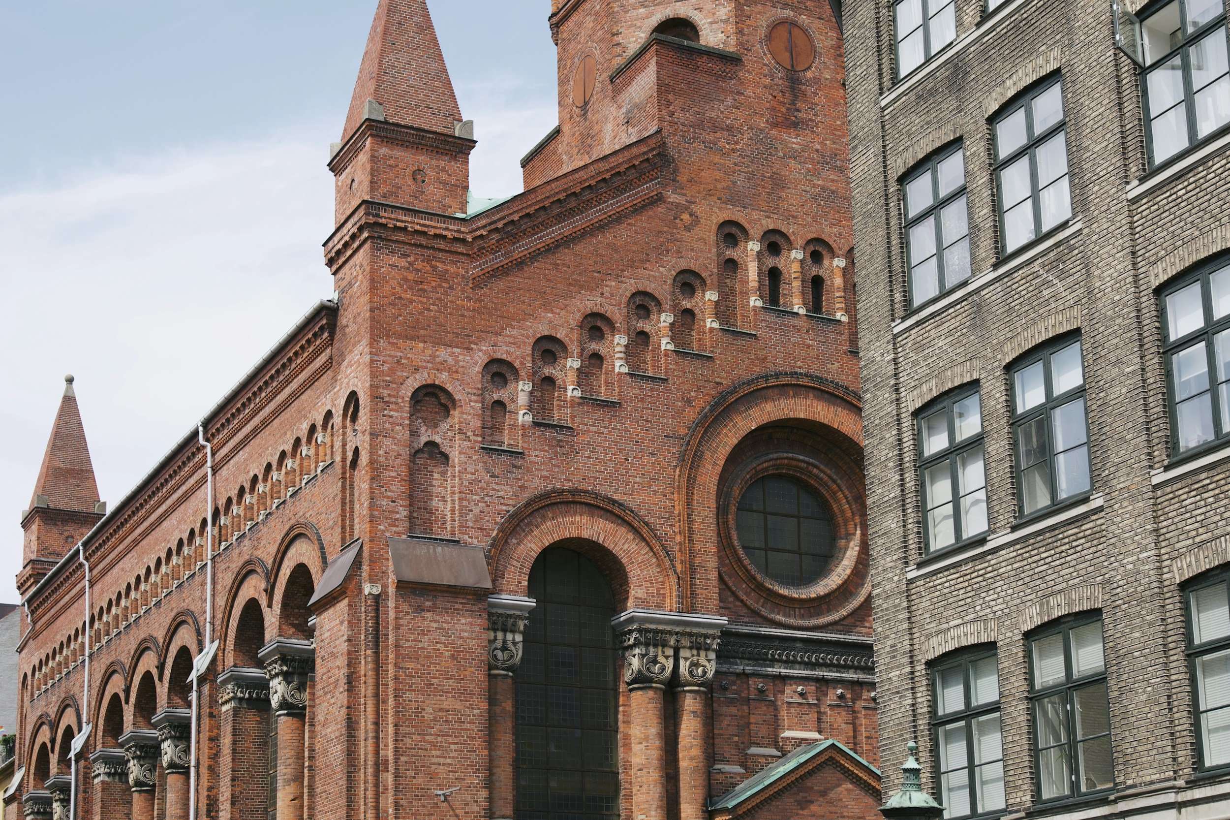 Close-up of historic red brick church with arched windows, circular window, tower, and decorative brickwork, with adjacent multi-story brick building with large rectangular windows.