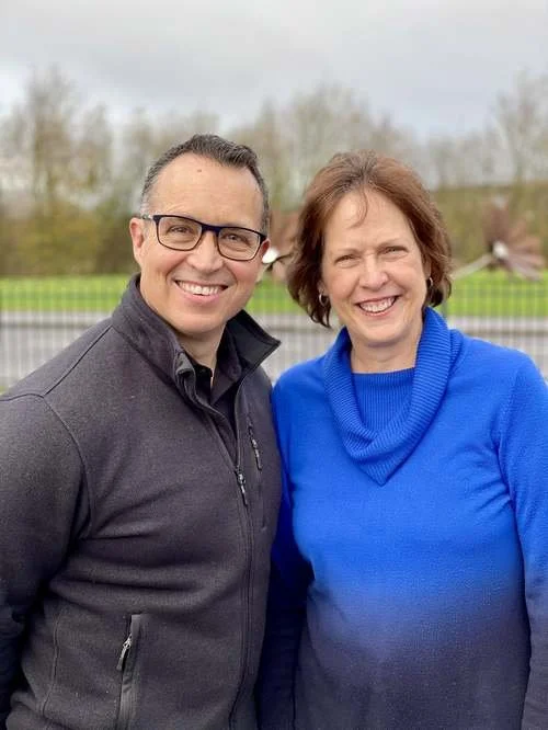 A man and a woman standing outdoors, smiling at the camera, with trees and a fence in the background.