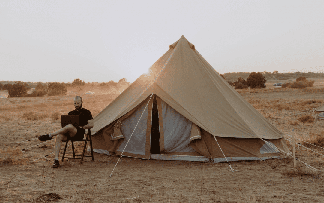 A man sitting on a chair using a laptop next to a large beige camping tent in an open, dry landscape during sunset.