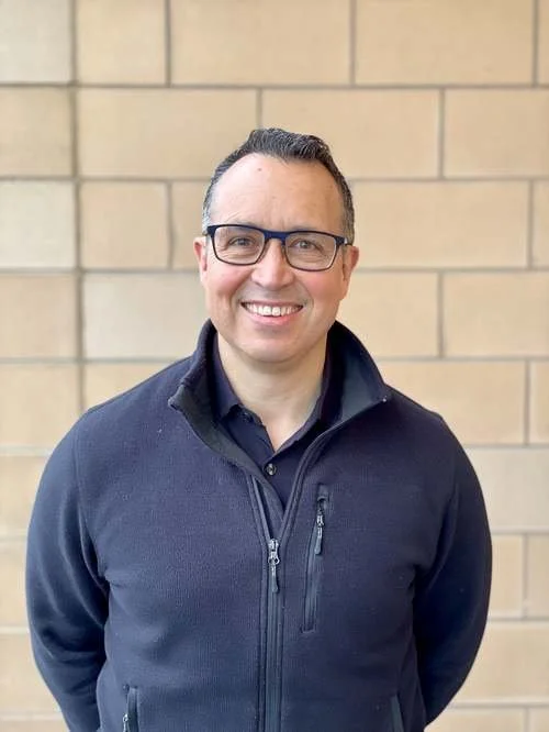 Smiling man with glasses wearing a navy jacket in front of a brick wall