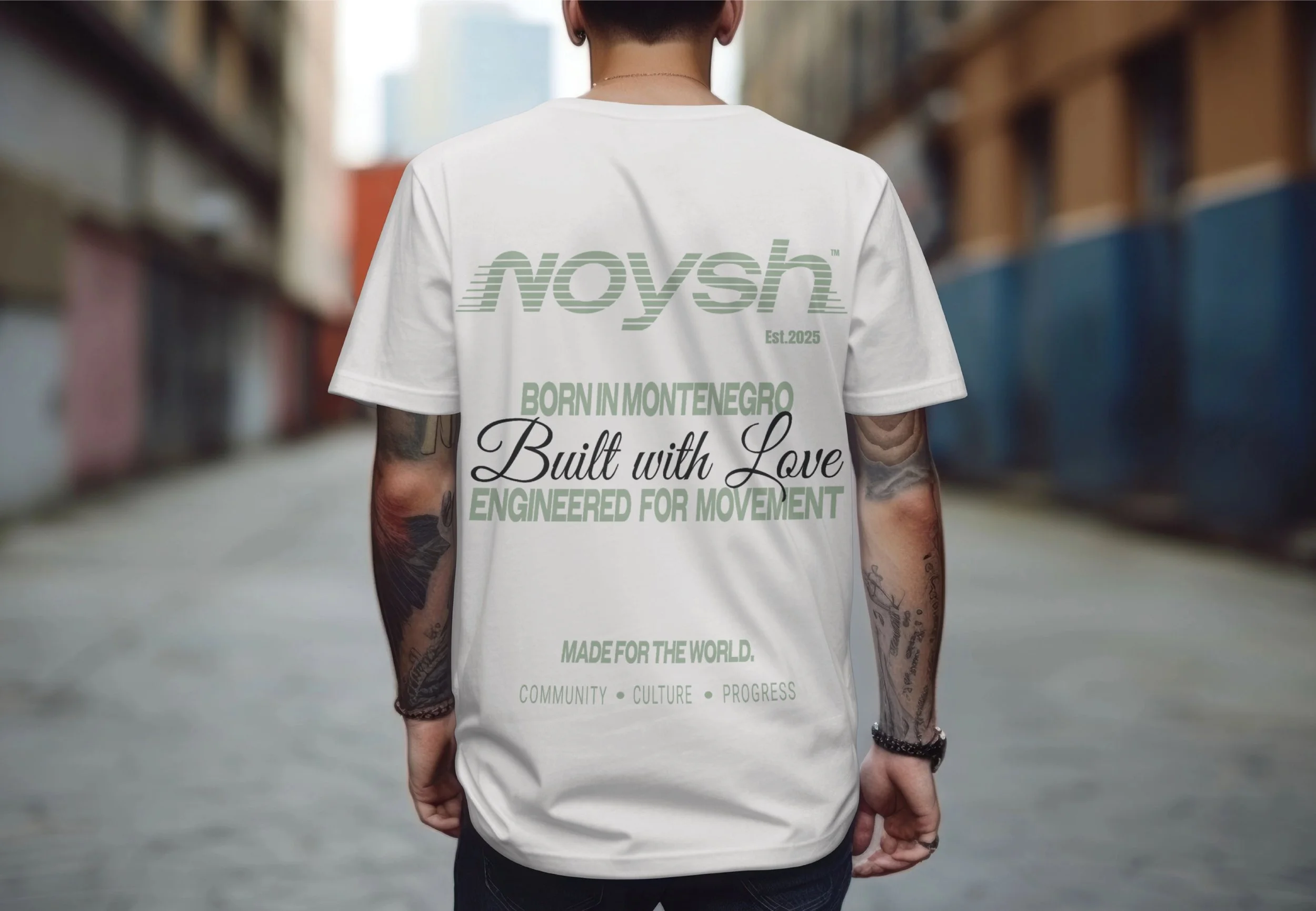 Back of a person wearing a white T-shirt with green and black text, standing on an urban street with blurred buildings on either side.