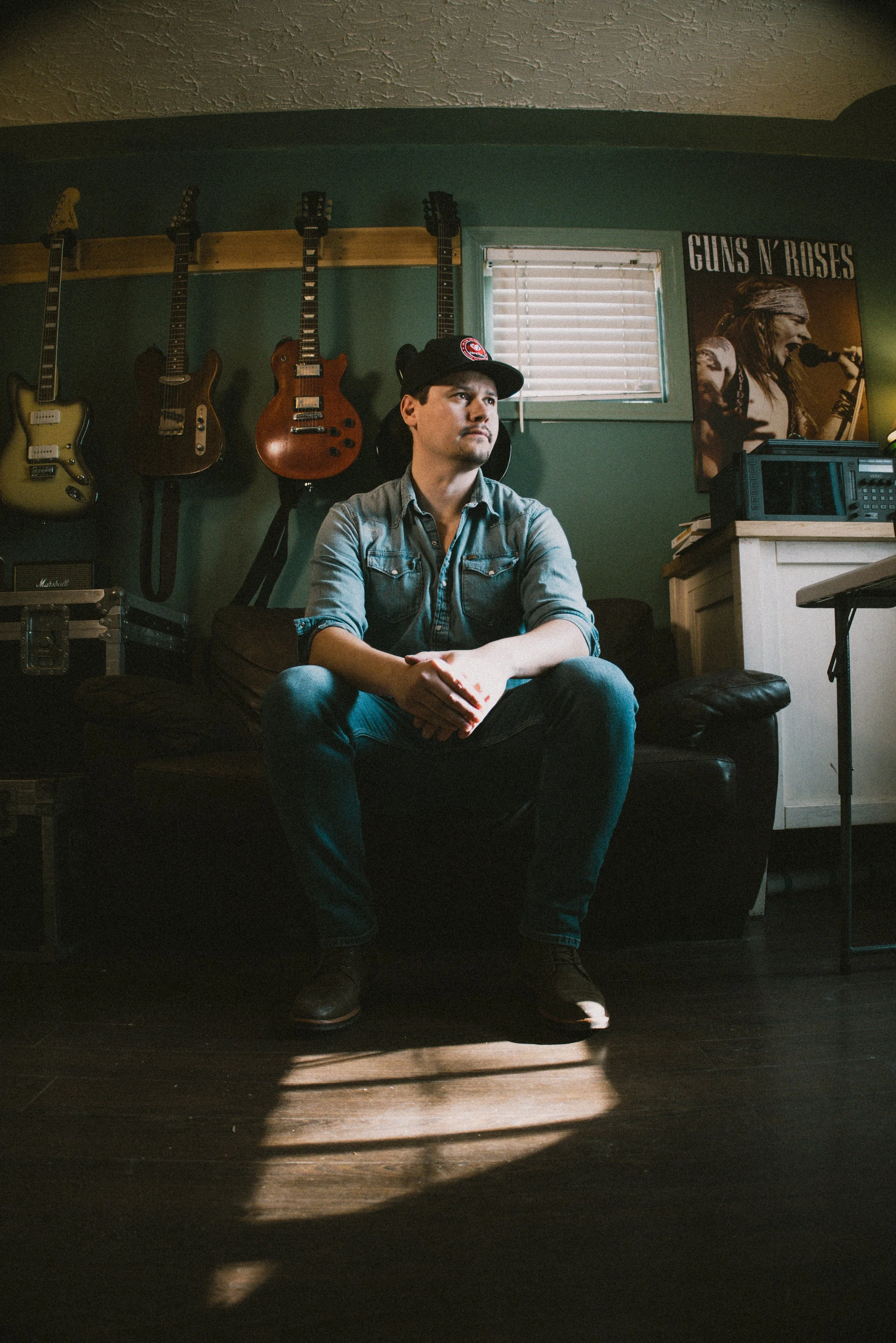 A man sitting on a black couch in a room with green walls and a shelf holding several guitars. A poster of Guns N' Roses is on the wall, and sunlight streams through a small window with horizontal blinds, creating a pattern on the dark wooden floor.