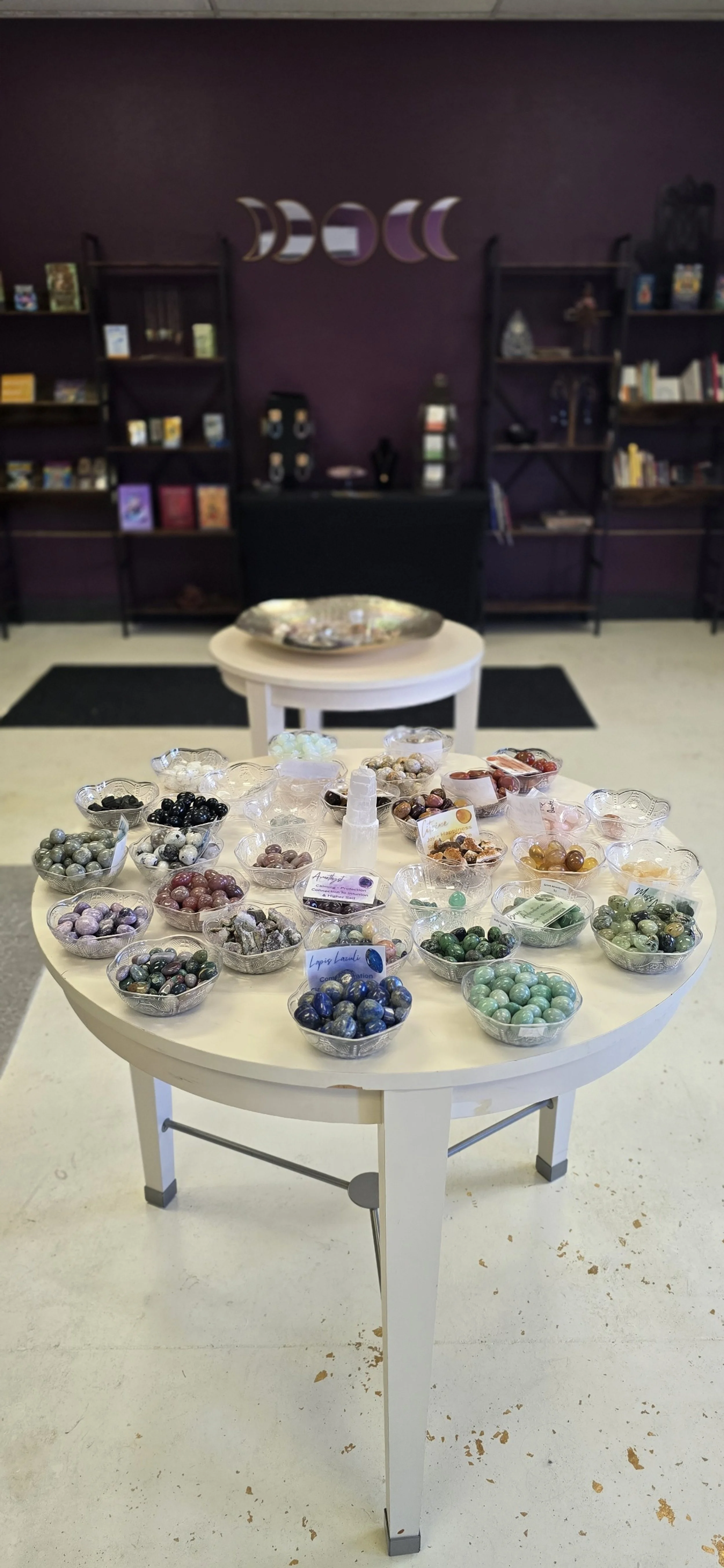 A white round table displaying an assortment of polished and tumbled crystals and stones in small glass bowls, with some labels and informational cards. In the background, there are black bookshelves filled with books and decorative items, and a purple wall with moon phase decor.
