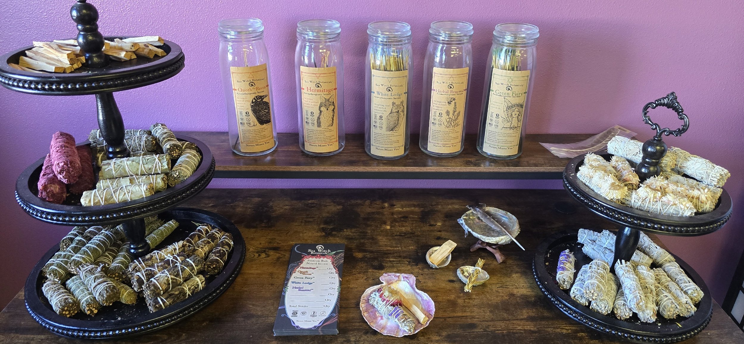 Display of various herbal incense sticks arranged on tiered trays and jars, with a pink wall background.